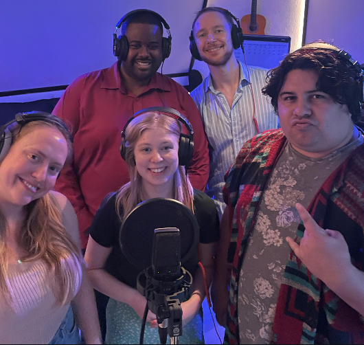 Group of five people in a recording studio, wearing headphones and smiling, with a microphone in front of them.