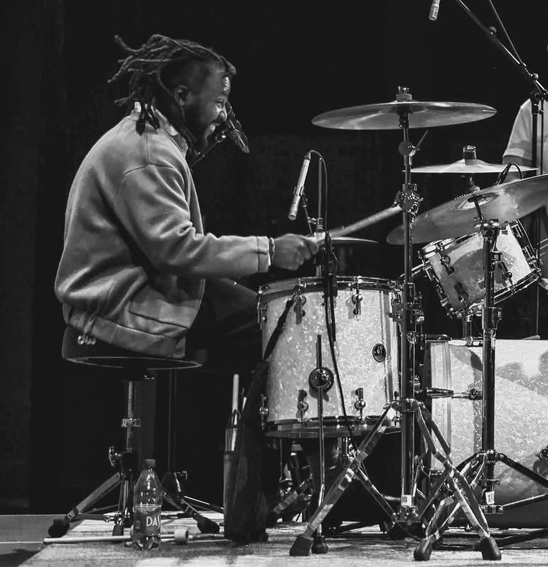 A man with dreadlocks playing the drums on stage, with a focused expression, surrounded by drums and cymbals, in a black-and-white photo.