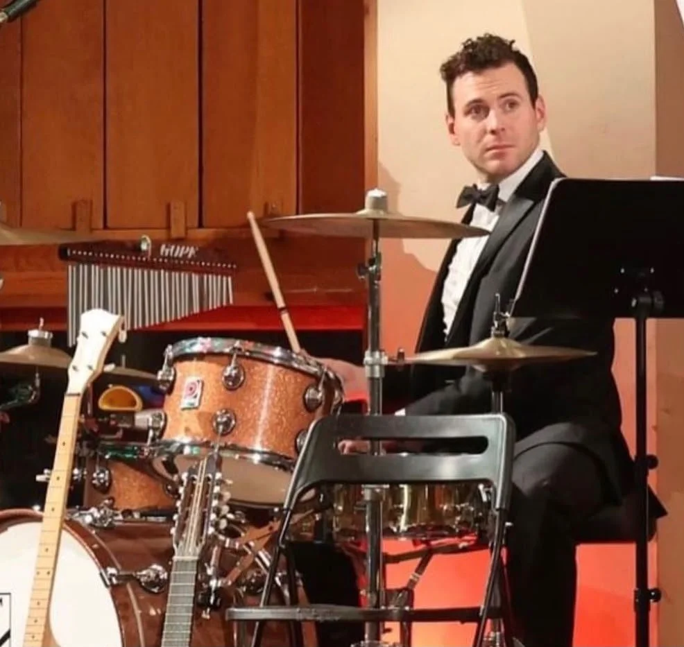 Man in a tuxedo sitting at a drum set, with a guitar and music stand nearby, in a performance setting.