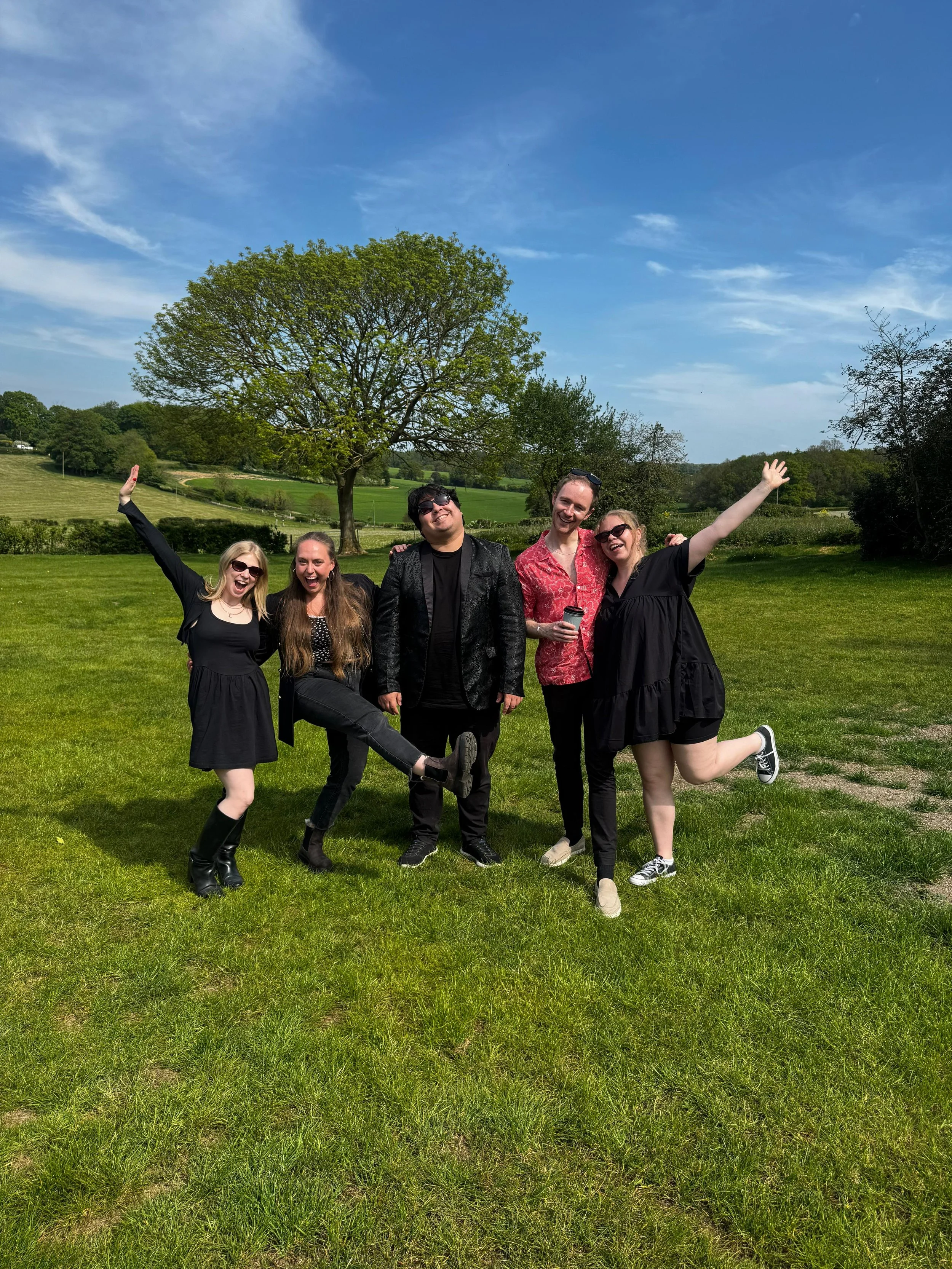 Group of five young adults smiling and posing happily on a green grassy field with trees and a blue sky in the background.