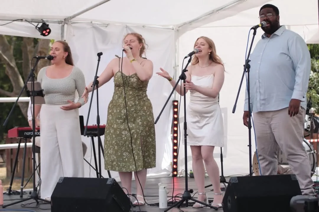 Four singers performing on stage inside a white tent, with keyboards and microphones, during an outdoor event.