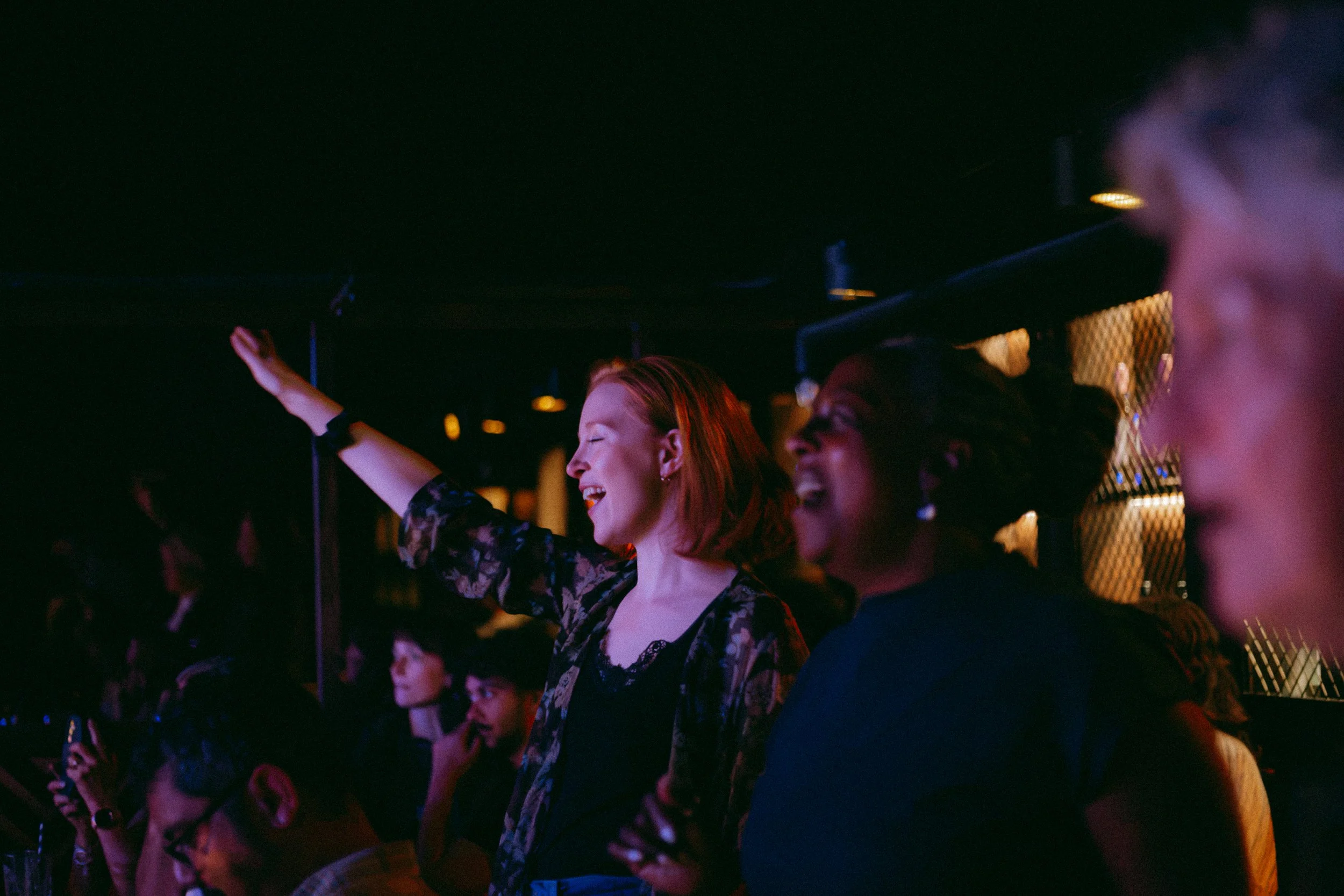 Group of five musicians performing on stage, with a woman singing into a microphone and others playing guitar and bass, in a dimly lit venue with audience watching.
