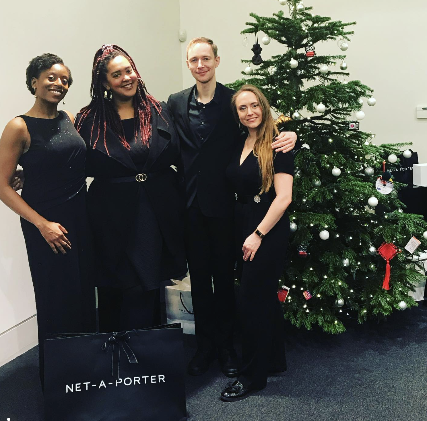 Four people standing next to a decorated Christmas tree, dressed in formal black attire, smiling at a holiday gathering.