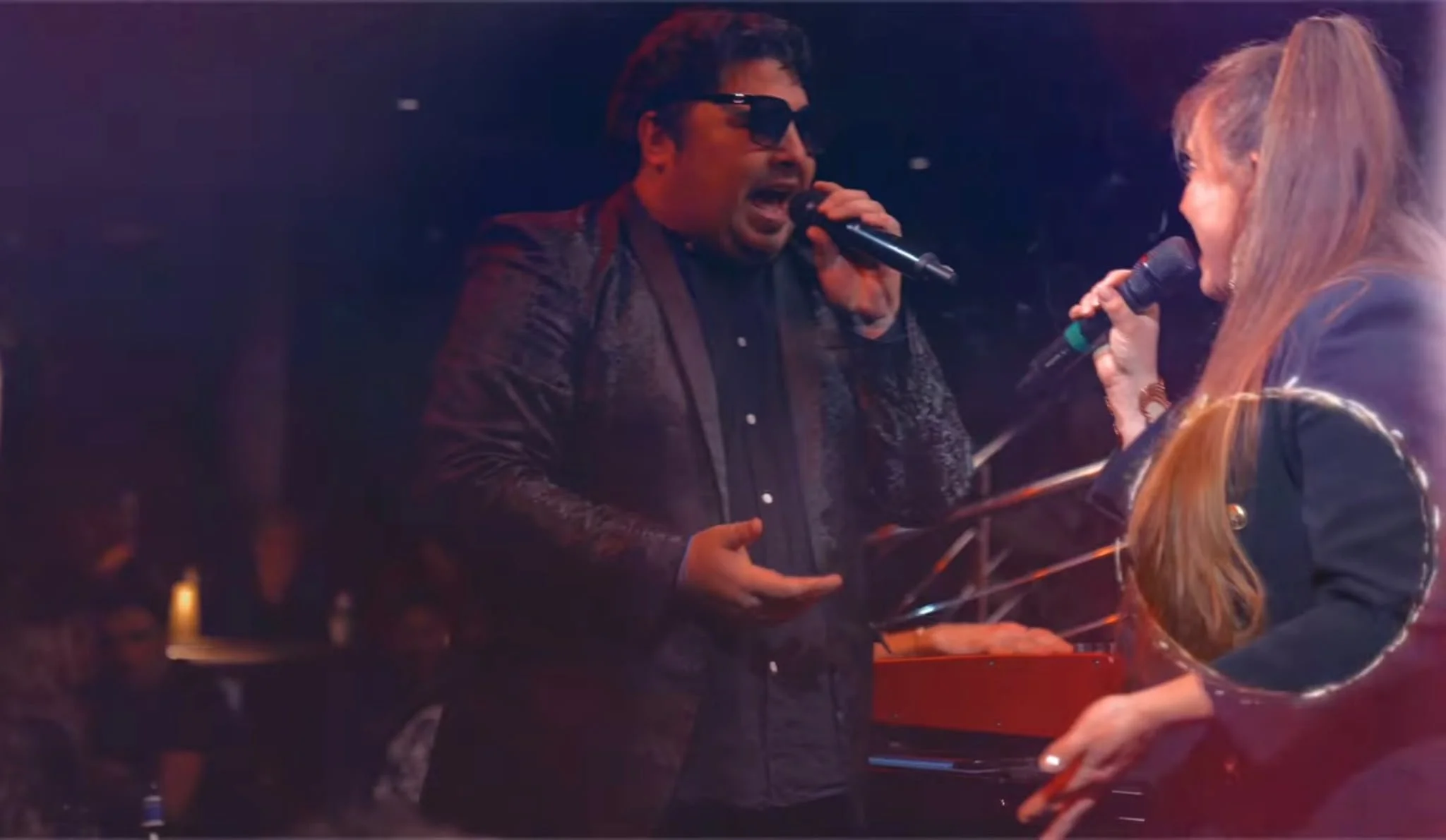 A man and woman singing into microphones on stage, illuminated by colourful lights.
