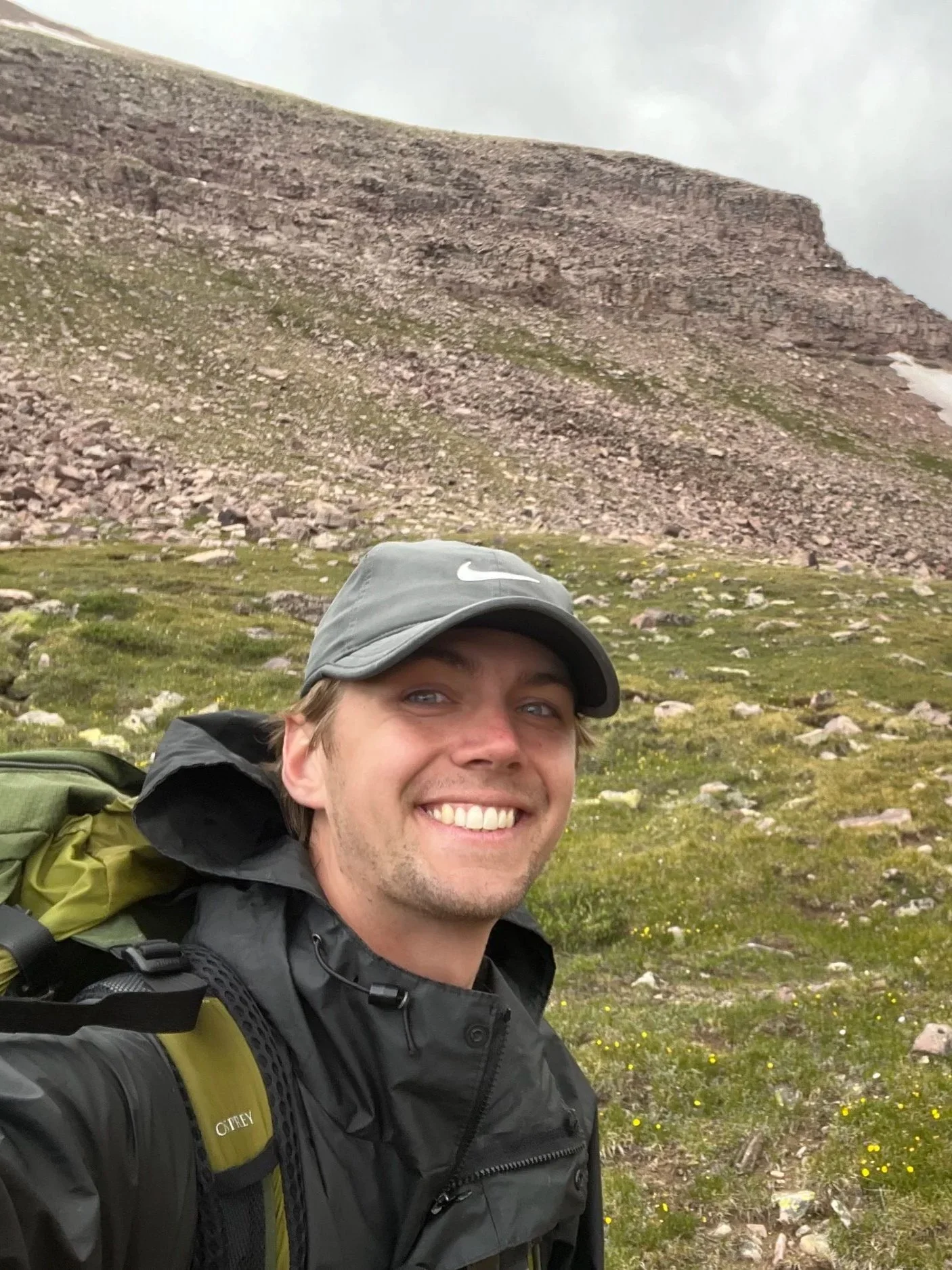 A smiling man wearing a gray Nike cap and a black jacket with a green hiking backpack taking a selfie in a mountainous outdoor setting with rocky terrain and patches of green grass.