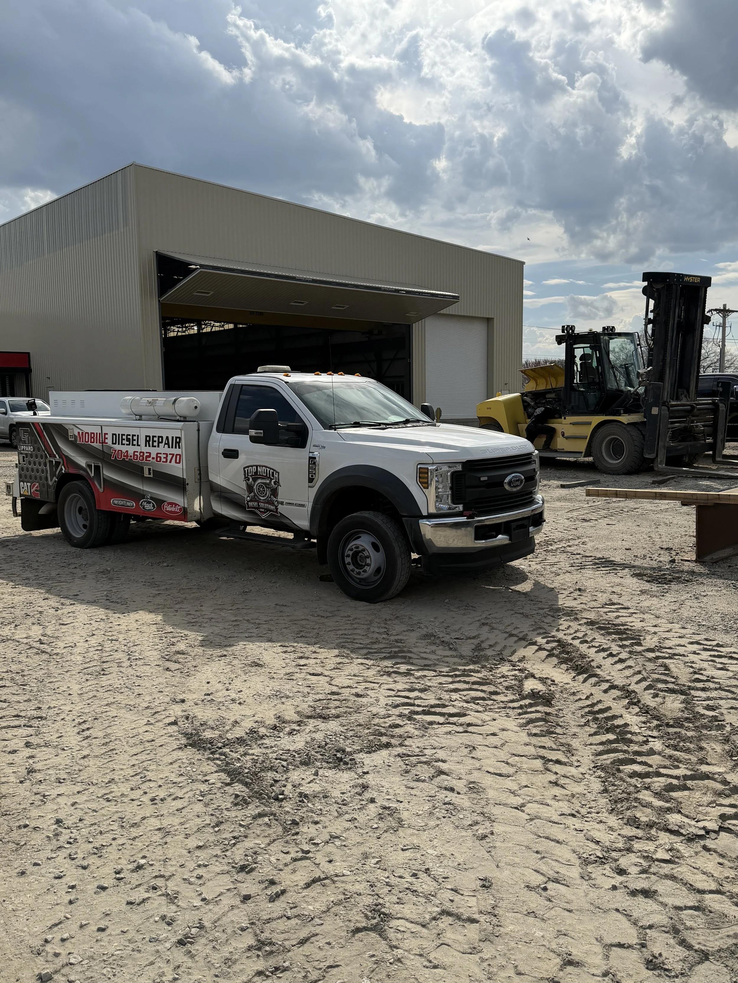 A construction yard with a white Ford truck from Top Notch Diesel Solutions Inc. and a yellow forklift in front of a large beige metal building, under a partly cloudy sky.