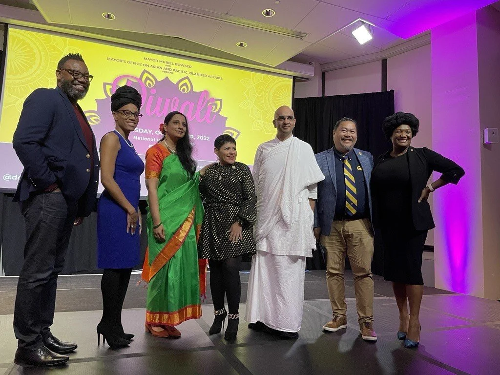 Group photo of Black man, Black woman, Southeast Asian woman, Latina woman, Southeast Asian man, Philippino man, and Black woman standing in front of Diwali poster on stage