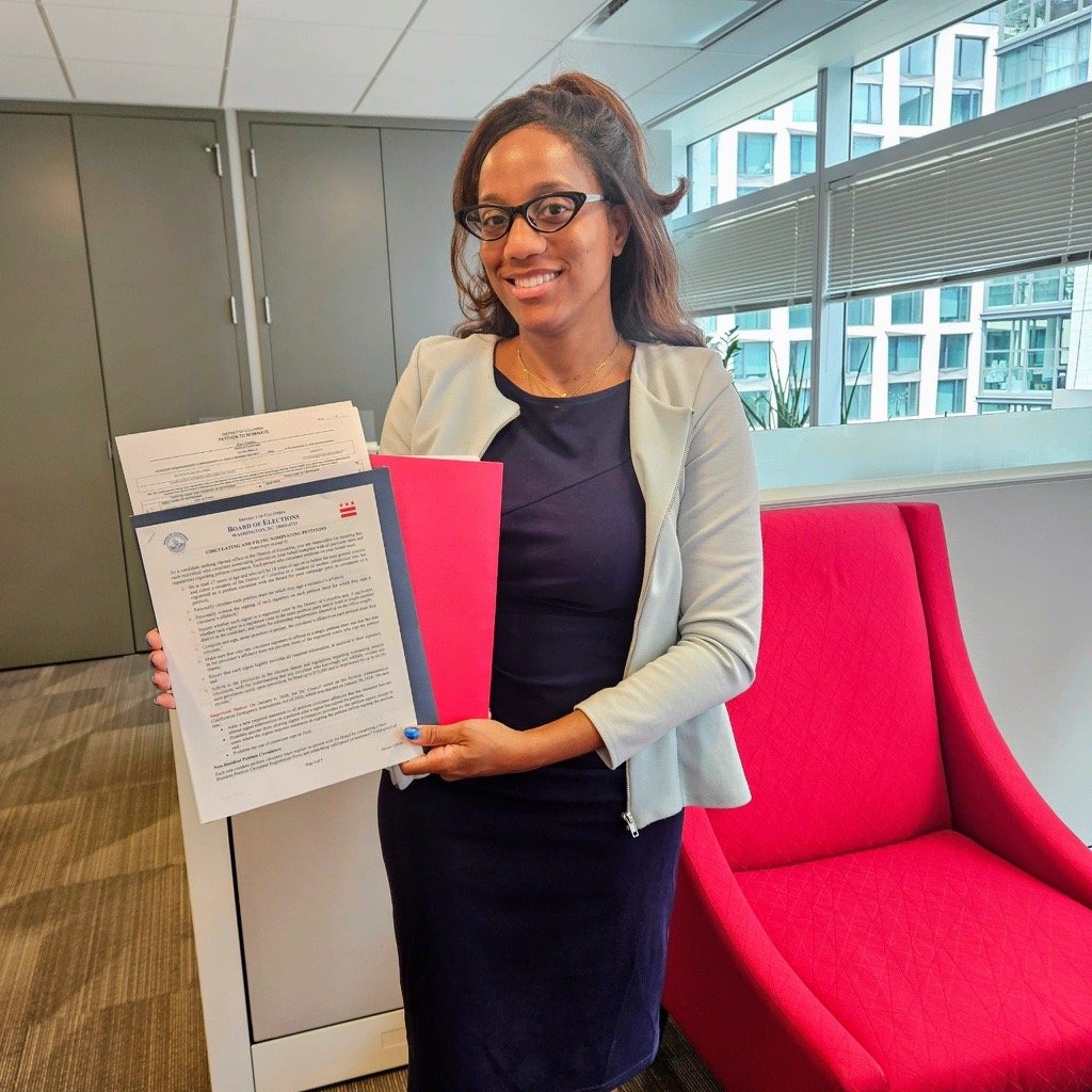 Black woman wearing blue dress and light blue blazer holding up her candidacy form with red folder background of the papers, she's in an office