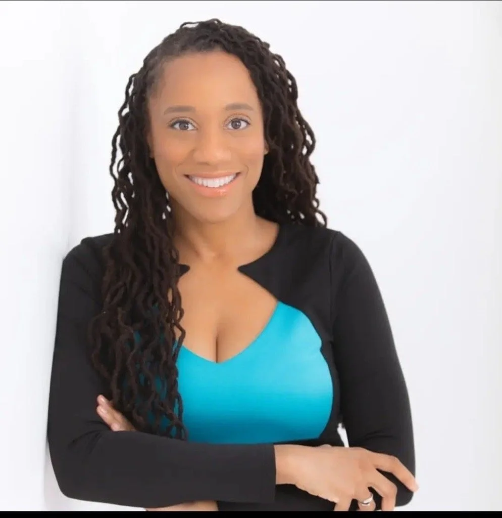 Headshot of Kari, Black woman with dreadlocks wearing black and teal with arms crossed and white background