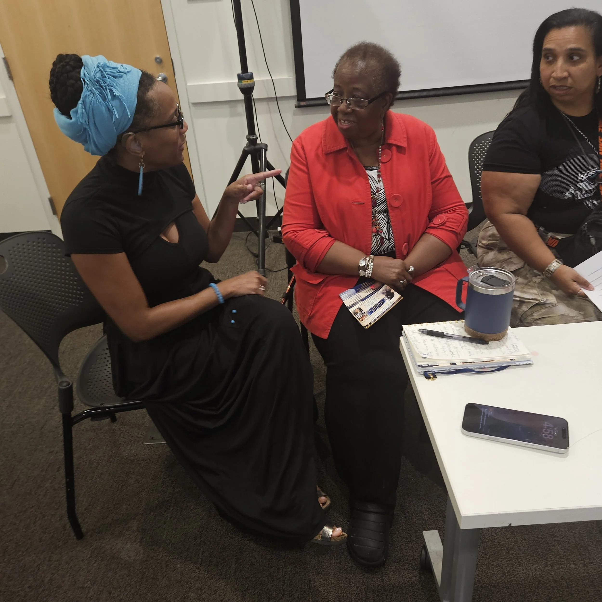 Picture of Black woman wearing black dress and light blue headscarf talking to two other Black women at a table. Image is in star shape