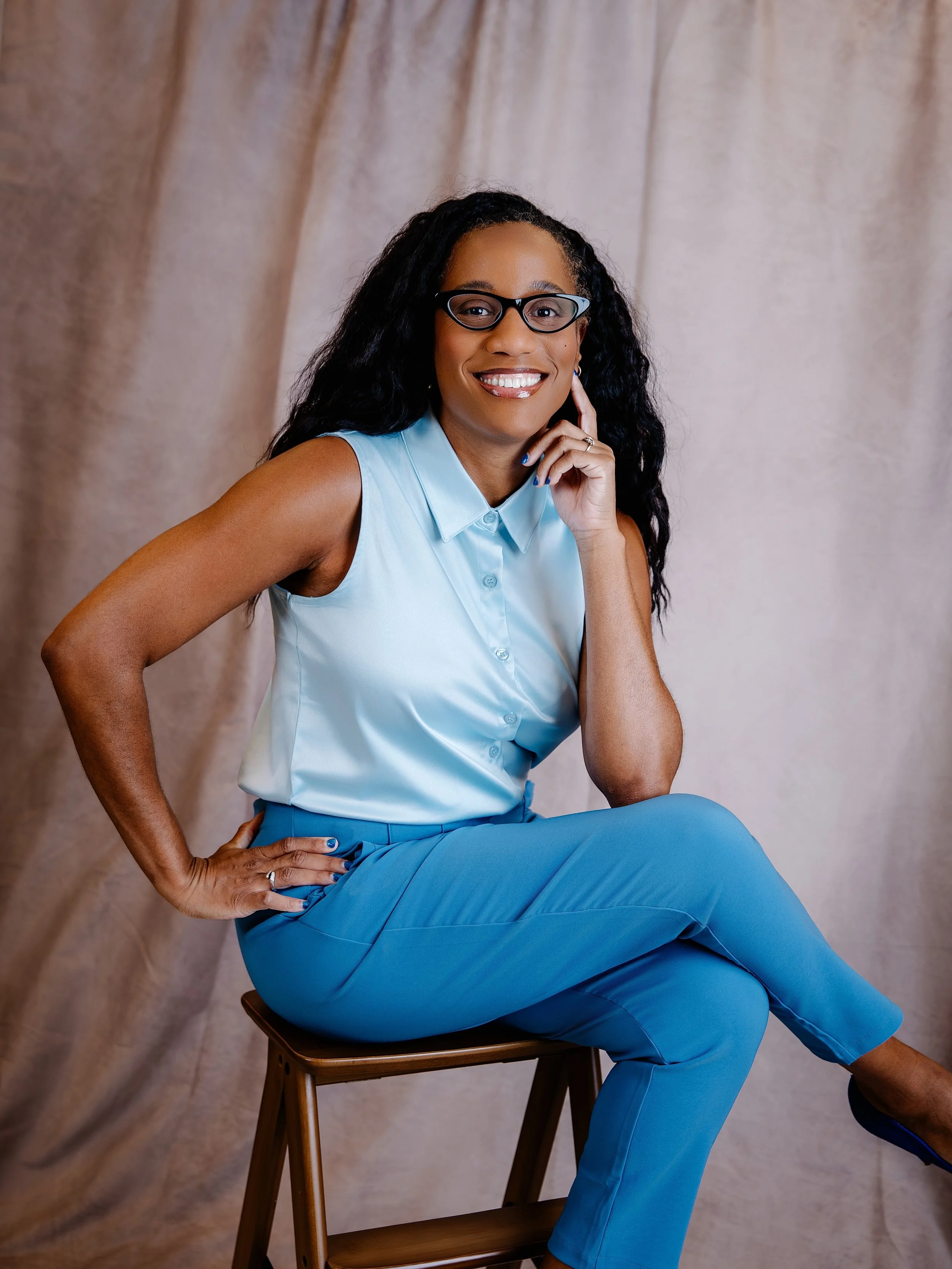 Black woman with hair down her back (blow dried thickness), wearing light blue sleeveless blouse and dusty blue pants. She's posed with elbow on knee and brown background