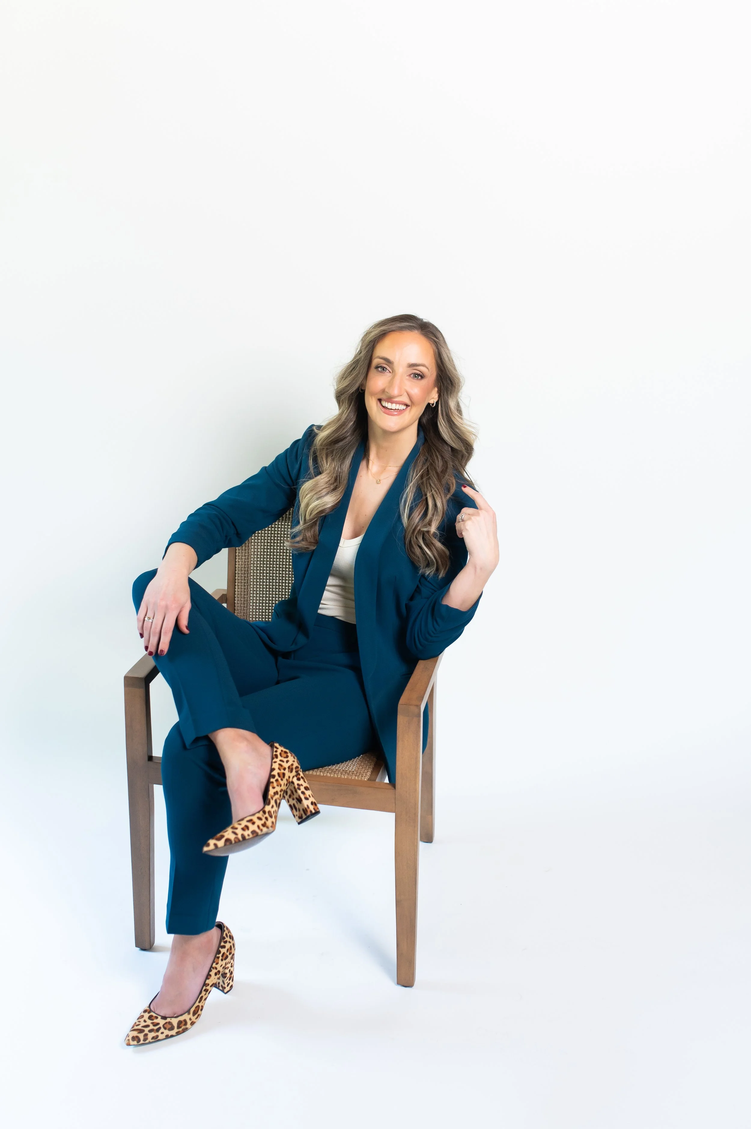 A woman with long wavy hair, smiling, sitting relaxed in a wooden chair against a plain white background. She is wearing a dark blue suit, leopard print high heels, and a white top.