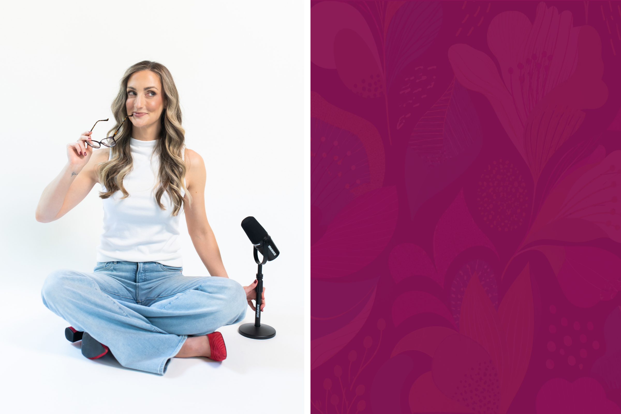A woman with long wavy hair, wearing a white sleeveless top and blue jeans, sitting cross-legged on the floor, holding glasses in her right hand, while a microphone on a stand is positioned in front of her.