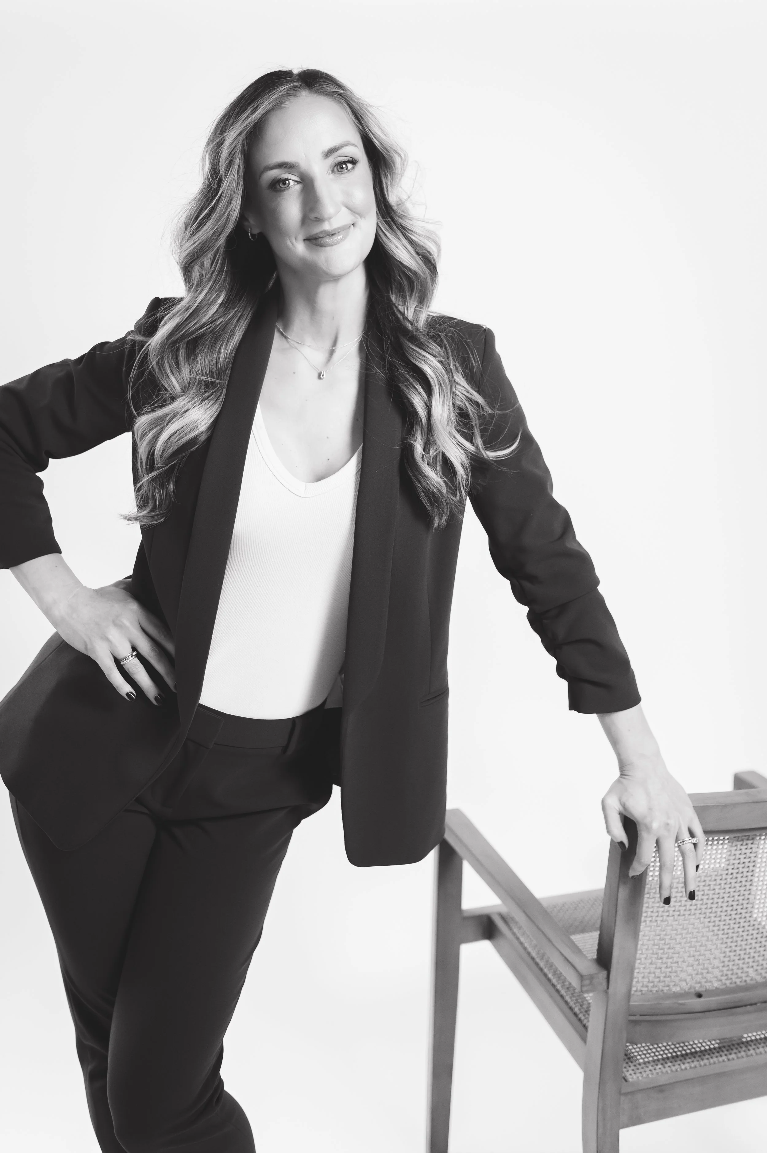Black and white photo of a woman with long wavy hair, wearing a blazer and pants, posing with one hand on her hip and the other resting on a wooden chair.