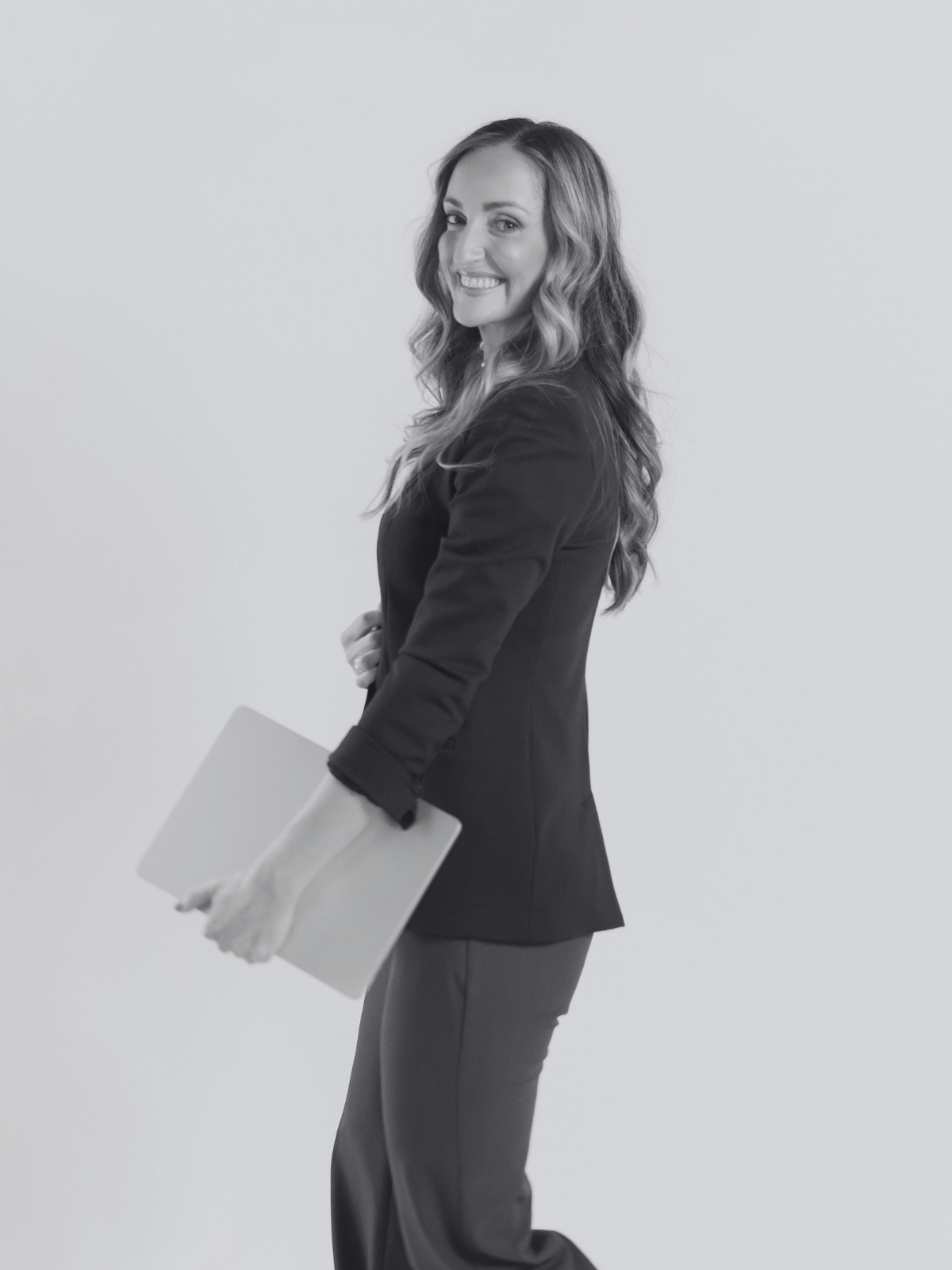 A woman with long wavy hair in business attire, holding a folder, smiling, standing against a plain background.