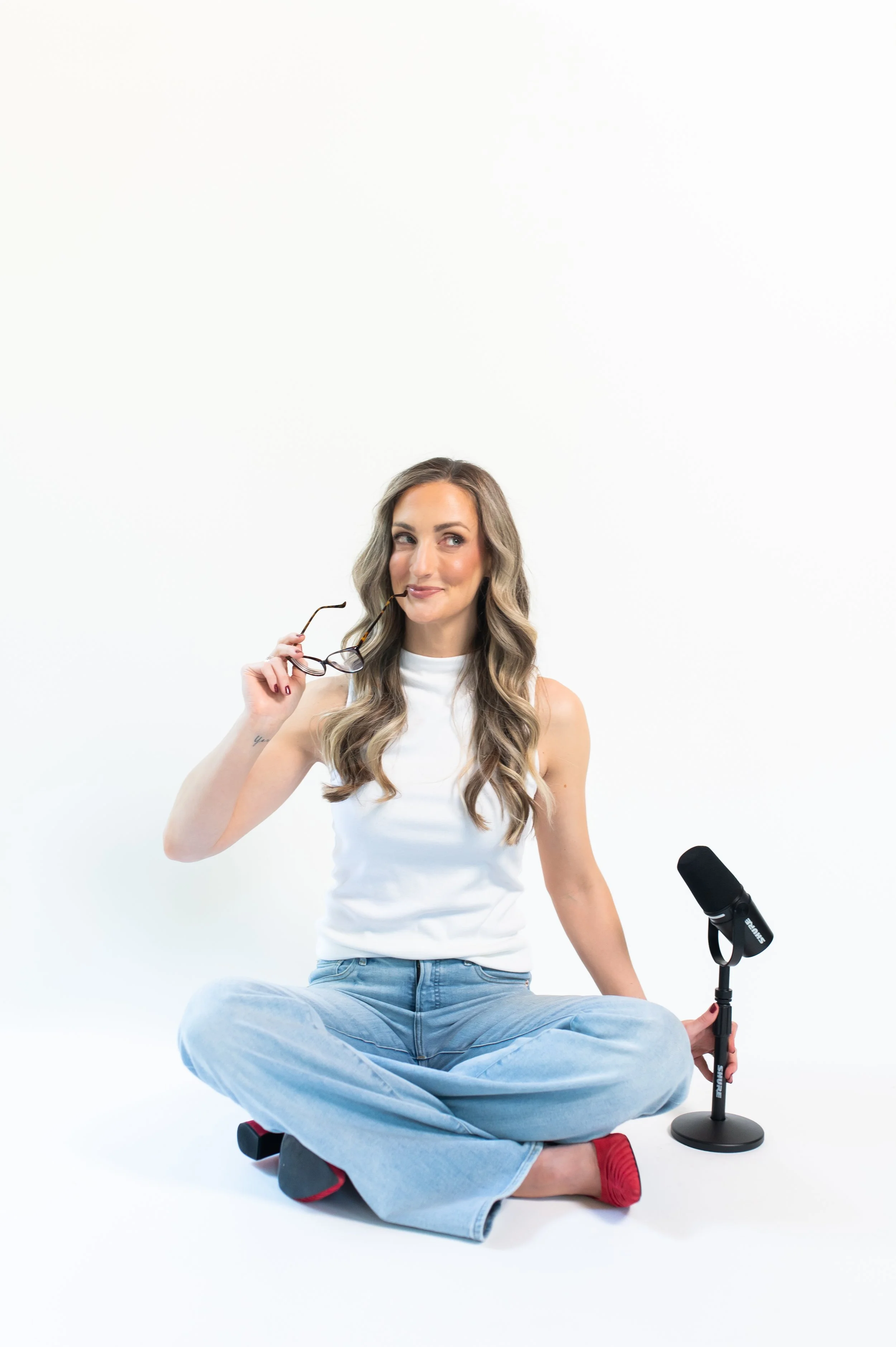 A woman with long wavy hair sitting cross-legged on the floor, holding glasses in one hand, with a microphone and a portable light stand nearby, against a plain white background.