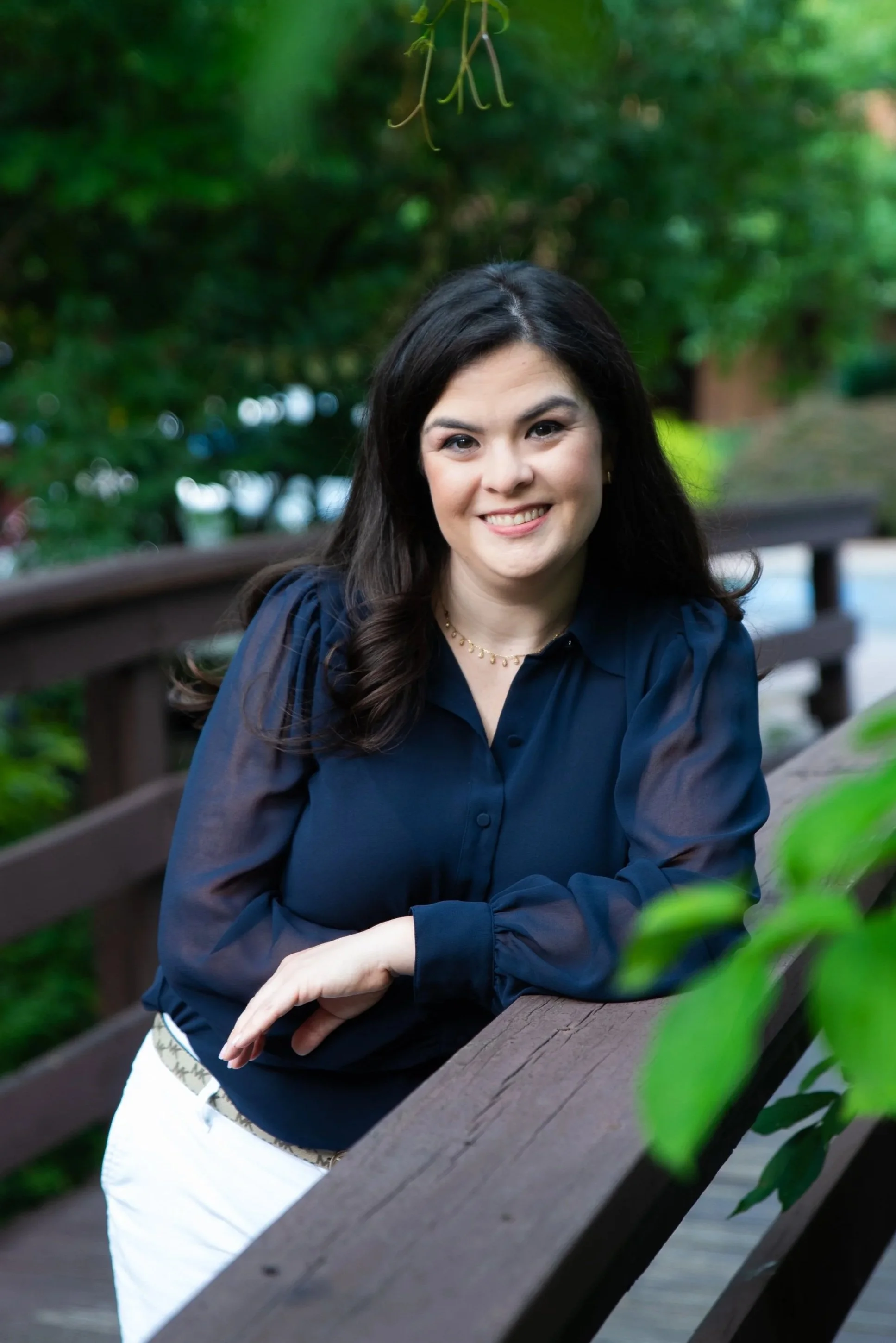 A young woman with long dark hair and a friendly smile, wearing a navy blue blouse and a white skirt with a patterned belt, sitting outdoors on a wooden bench in a lush green park.