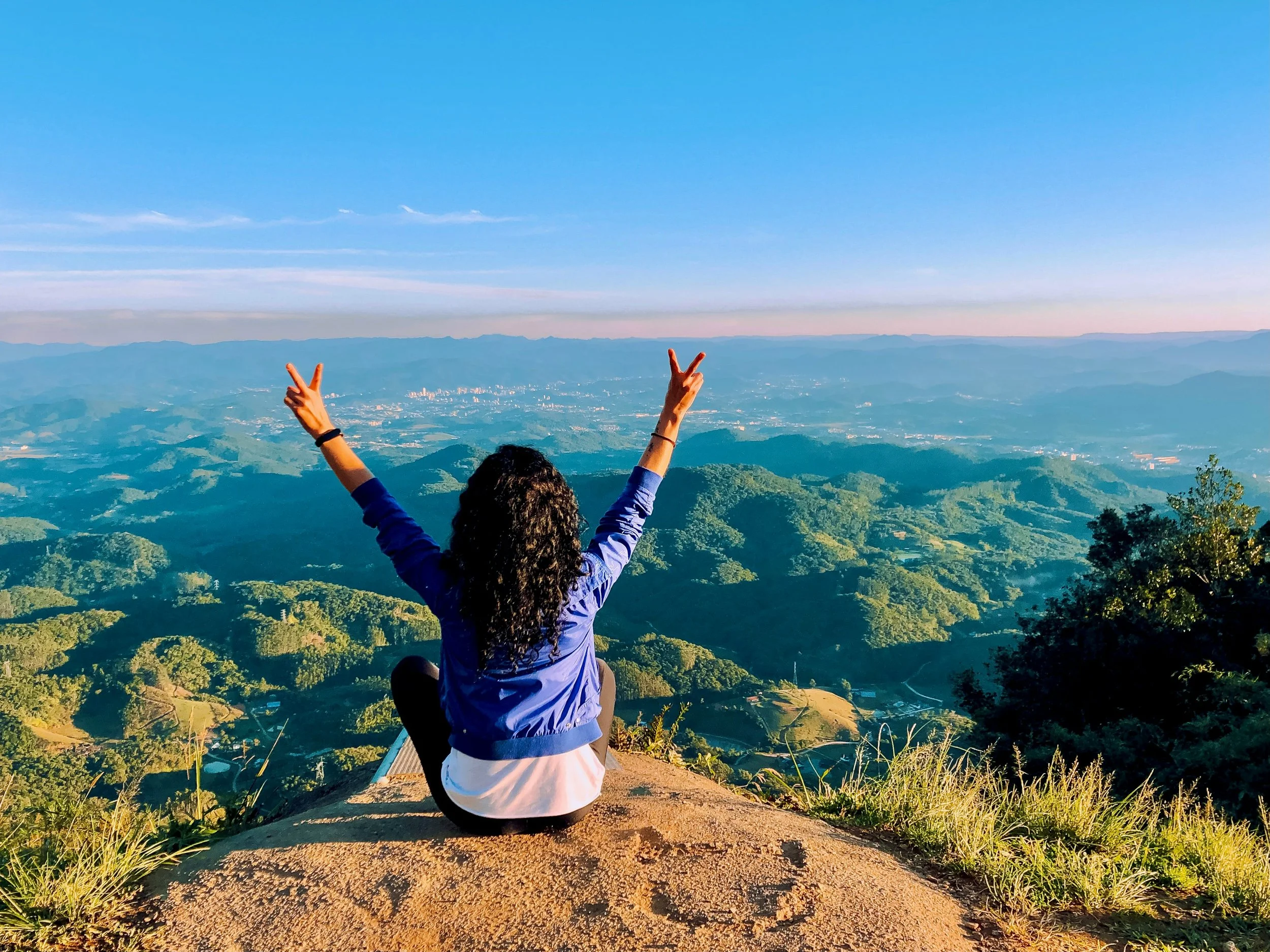 Woman on top of a mountain looking out over the valley below with her hands raised in peace signs