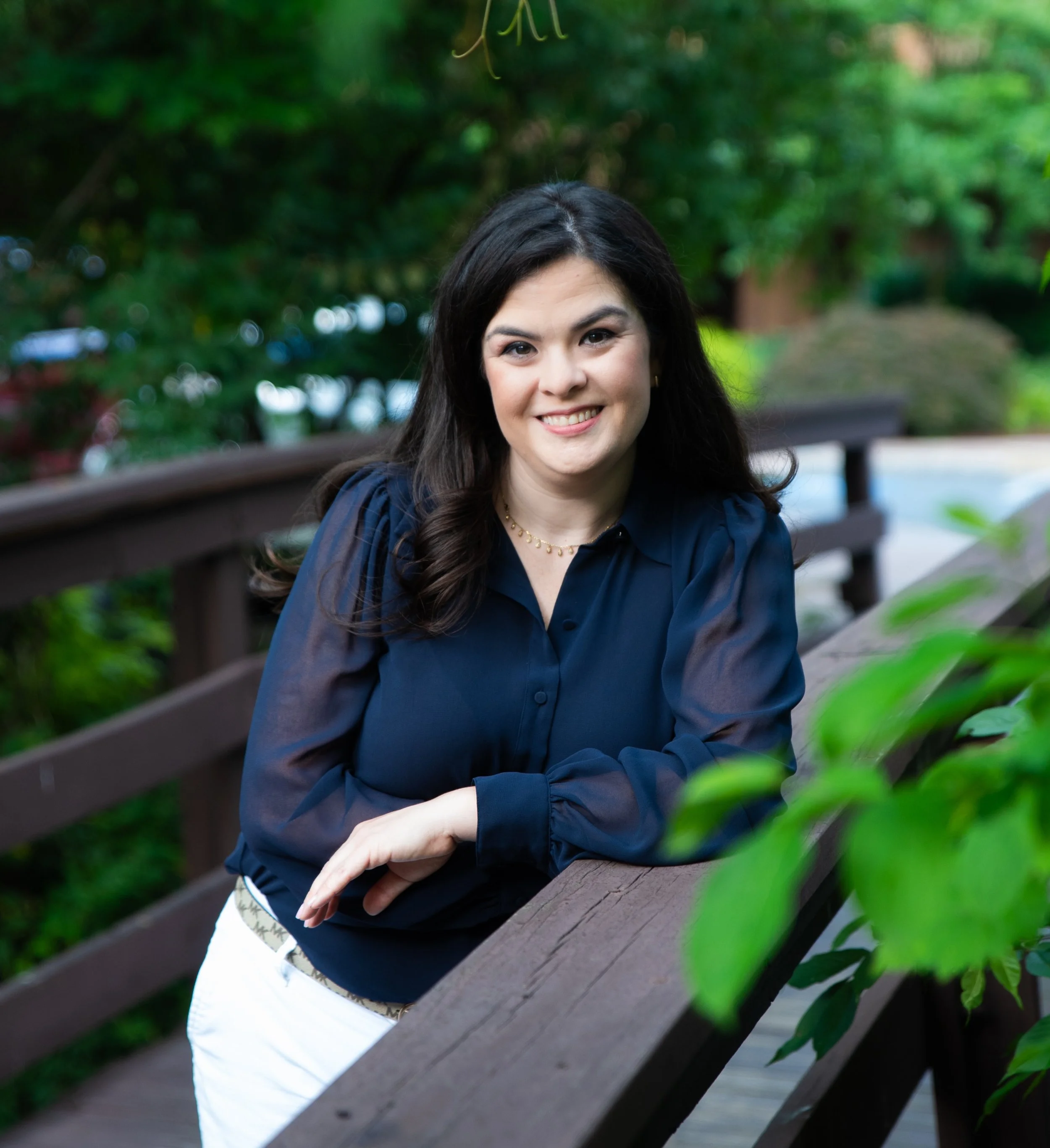 Brittany Vera, LCSW, a woman with dark brown hair wearing a navy blue blouse standing outside on a bridge surrounded by trees