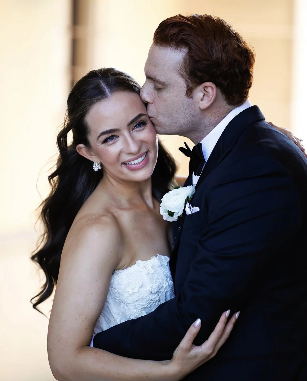 A bride and groom dancing at their wedding. The bride is smiling, wearing a strapless white wedding dress and pearl earrings. The groom, wearing a black tuxedo with a bow tie and boutonniere, is kissing her on the forehead.
