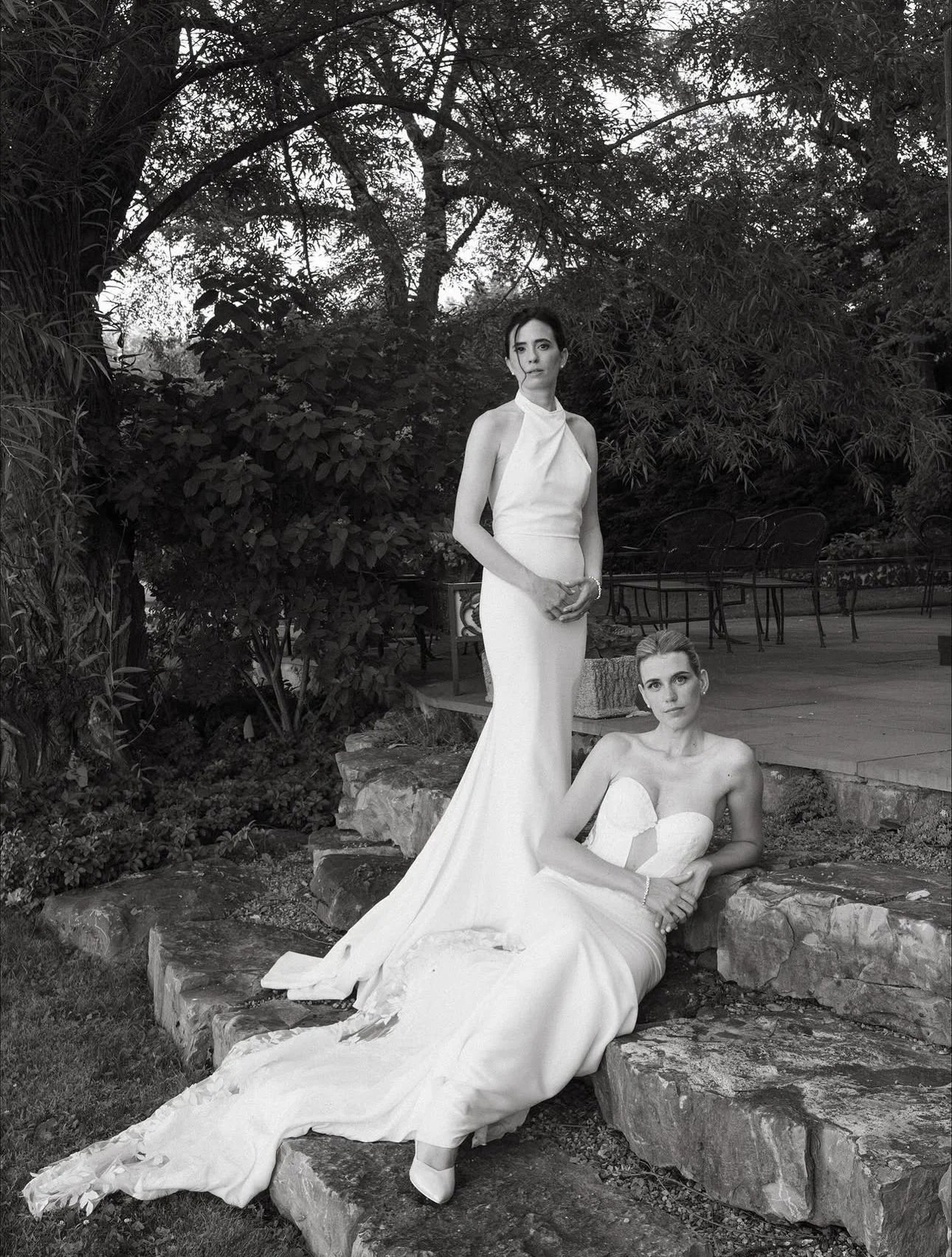 Two women in elegant white dresses outdoors among trees; one is standing and the other is sitting on rocks.
