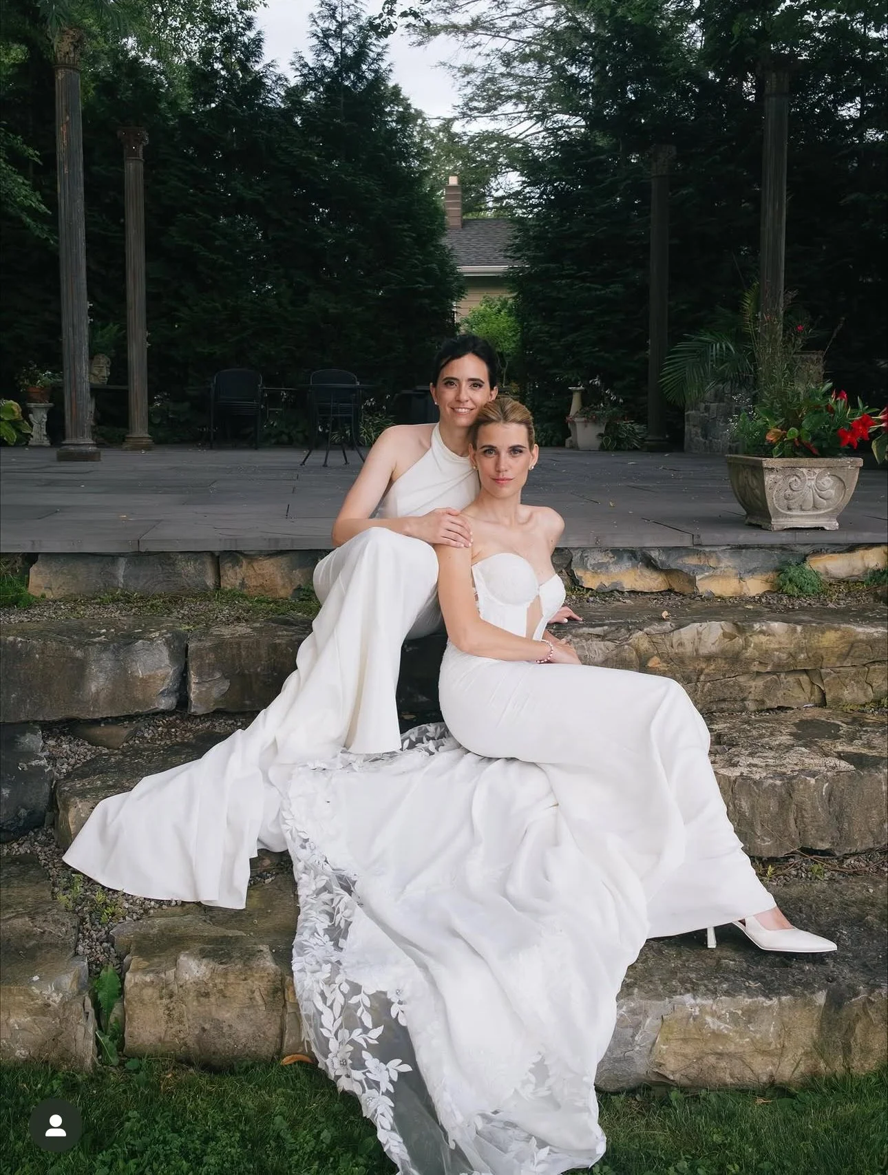 Two women dressed in white wedding gowns sitting on stone steps outdoors, surrounded by greenery and potted plants.