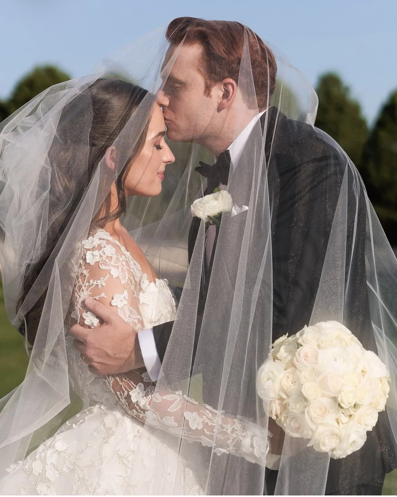 A bride and groom with a veil over both of them, kissing during their wedding outdoors. The bride is wearing a lace wedding gown, and the groom is in a tuxedo, holding a bouquet of white roses.