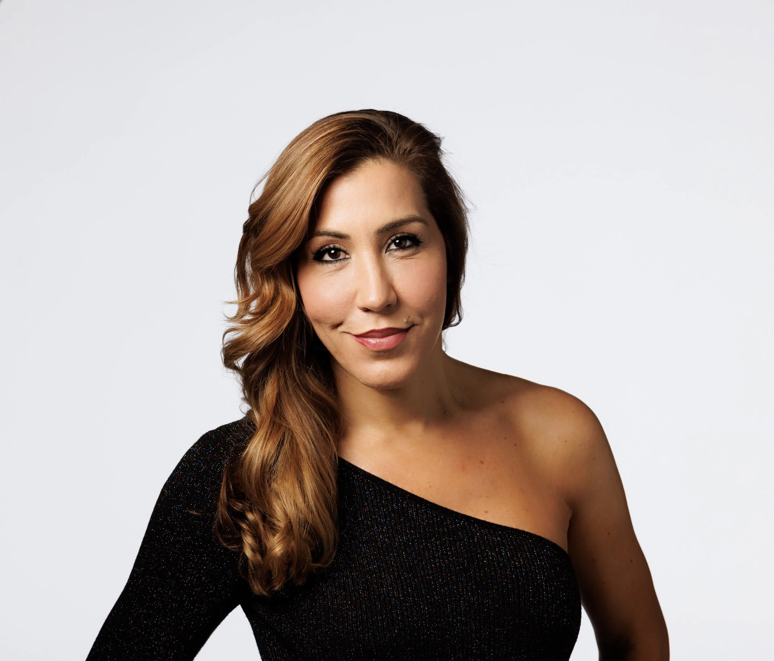 A woman with light skin and long, wavy light brown hair with highlights, wearing a black, off-shoulder, sparkly dress, smiling against a plain white background.