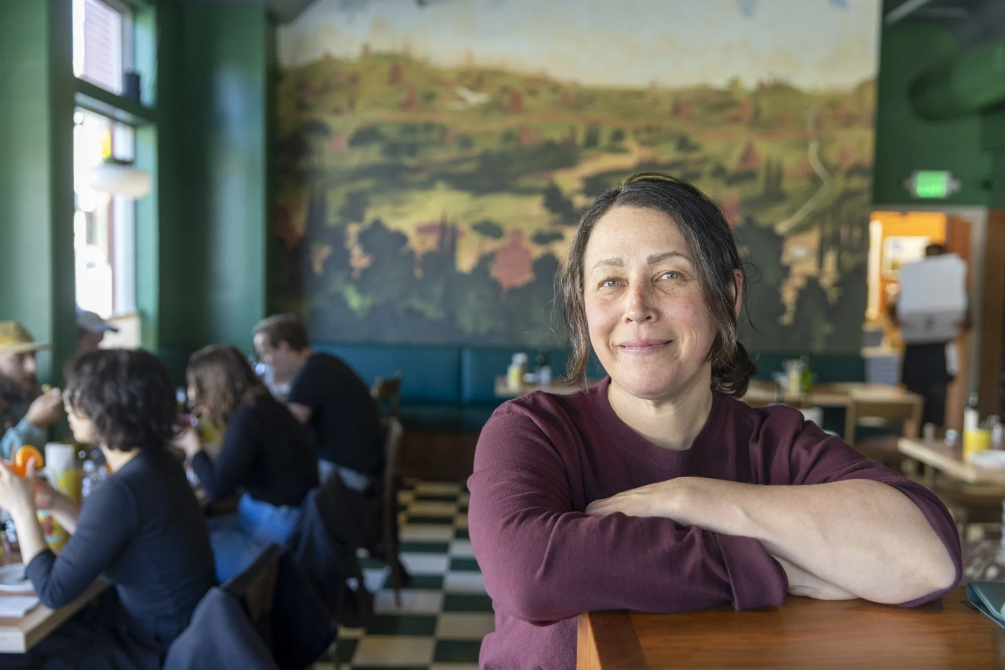 A woman with shoulder-length dark hair and a maroon shirt smiling at the camera while seated at a restaurant table. Behind her, other diners are eating, and a large scenic mural of hills and trees decorates the wall.