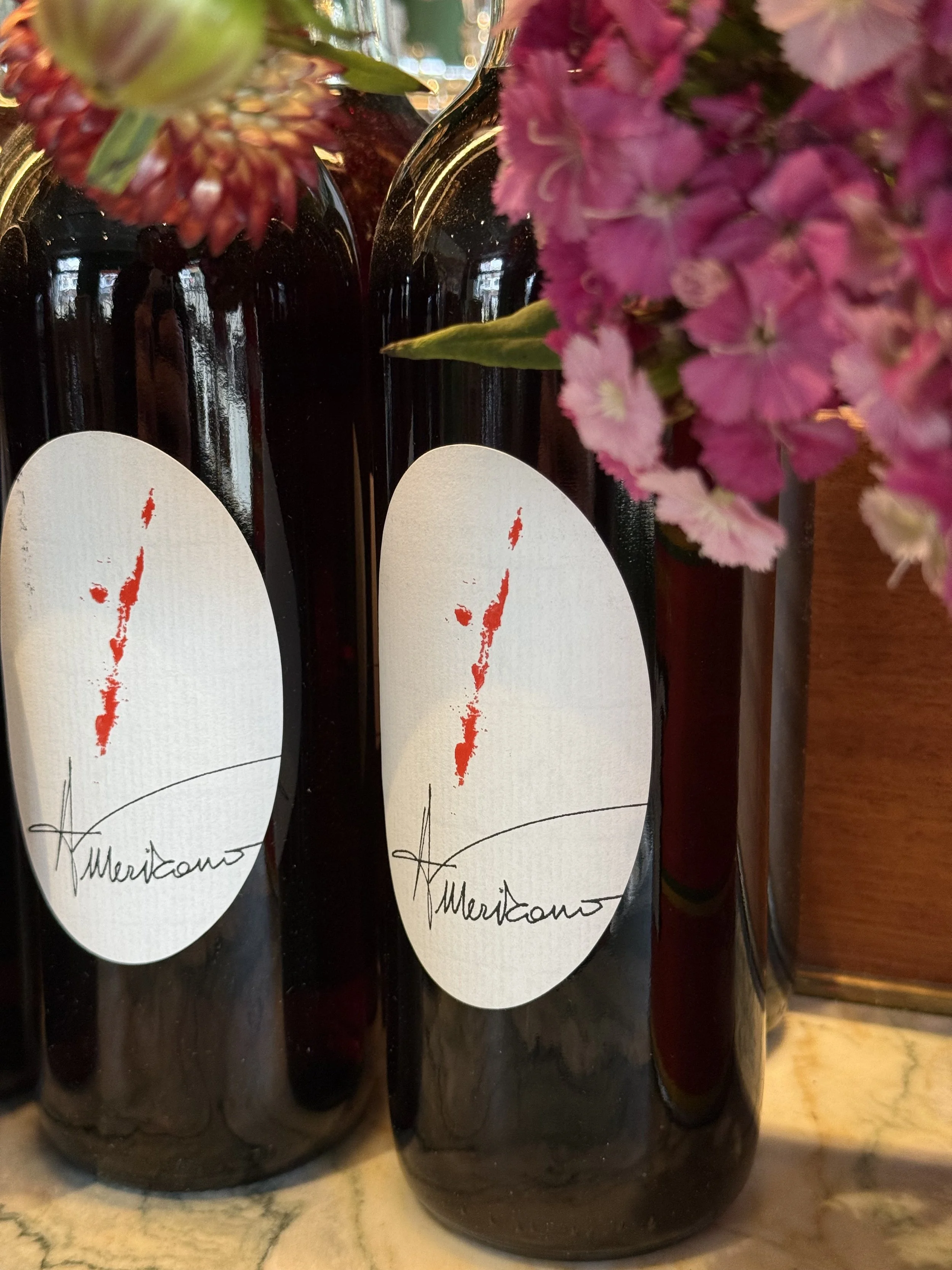 Three dark wine bottles with white labels and red abstract design, surrounded by pink and red flowers, placed on a marble surface.