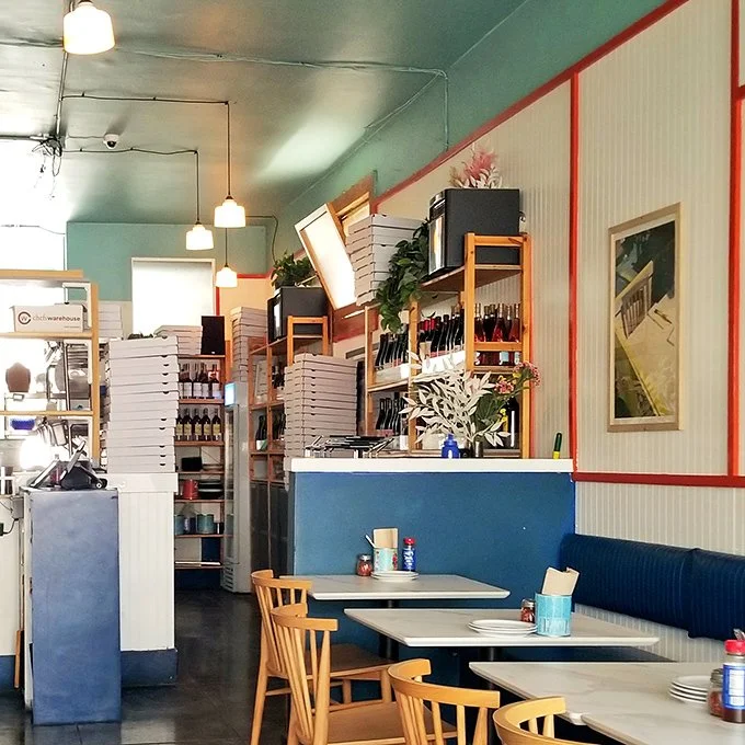 Interior of a restaurant with blue and white walls, wooden chairs, marble tables, and shelving filled with bottles and pizza boxes.