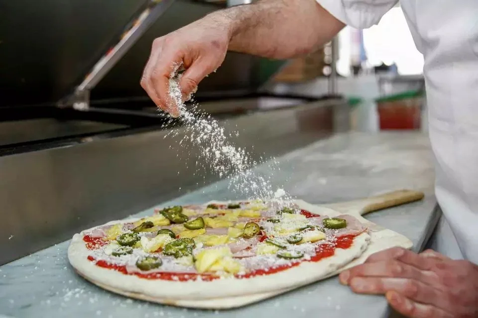Person sprinkling cheese onto a pizza topped with jalapeños, pineapple, ham, and cheese.