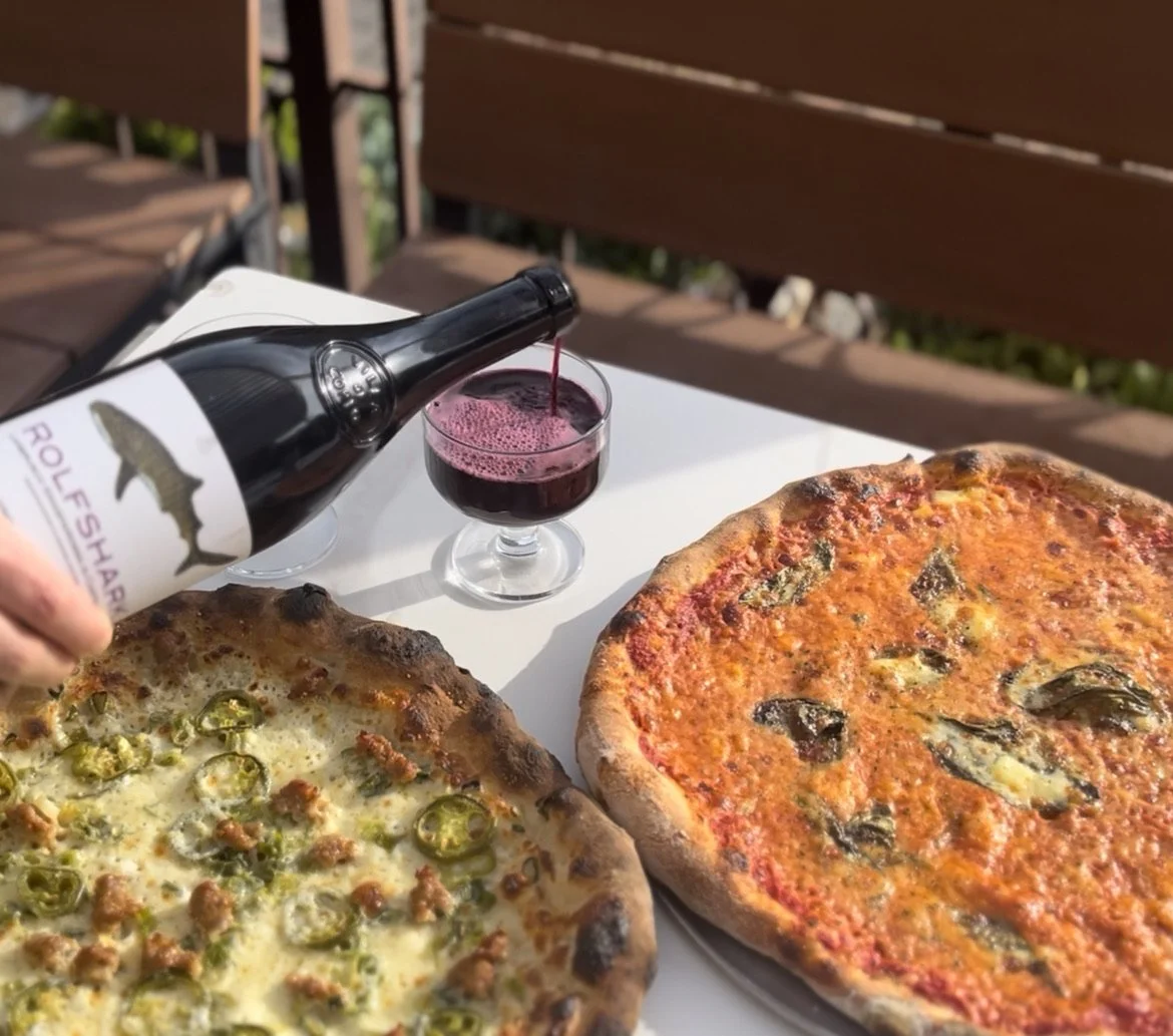 Pizza on a white table with a bottle of red wine being poured into a glass and a second glass already filled. Two pizzas, one with jalapeños and sausage, the other with basil and cheese, on a wooden bench outdoors.