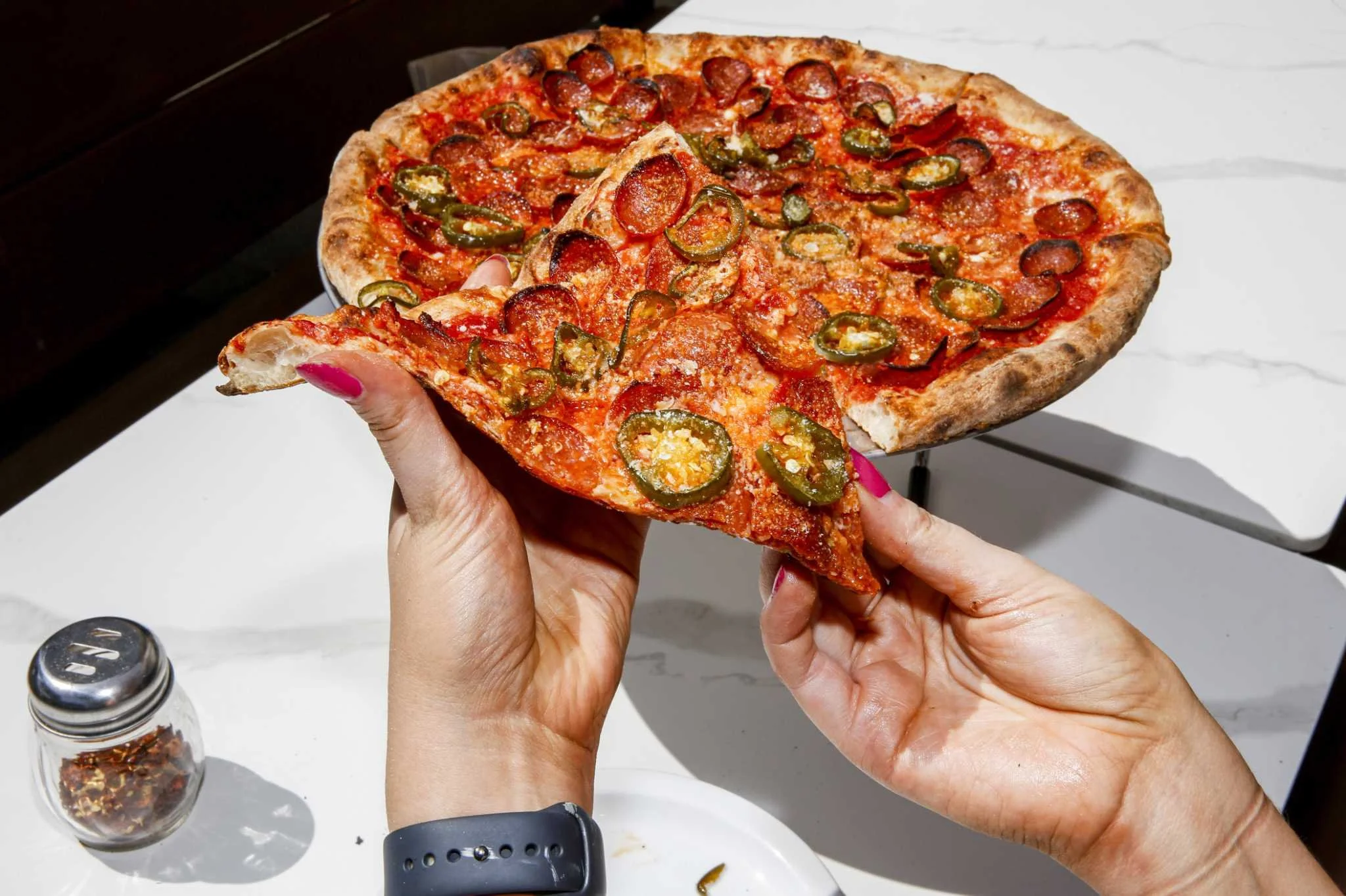 Close-up of a person holding a slice of pepperoni pizza with jalapeño slices on top, displaying a cheese and tomato sauce crust, with a white marble surface and a small jar of crushed red pepper flakes in the background.