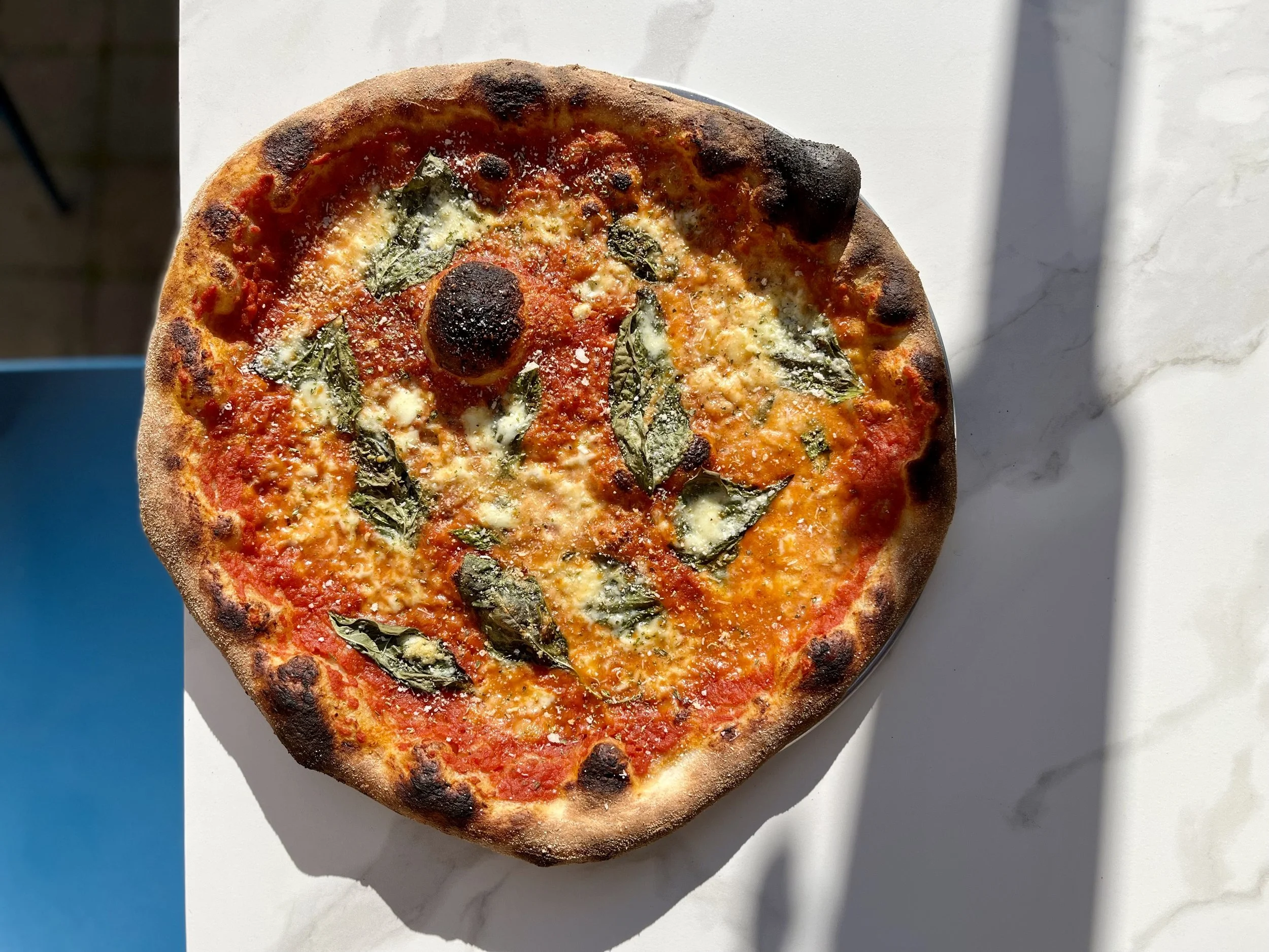 Close-up of a pizza with tomato sauce, mozzarella, basil leaves, and slightly charred crust, sitting on a white surface with a blue background.
