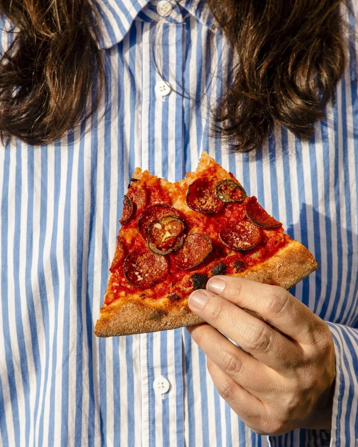 Person wearing a blue and white striped shirt holding a slice of pepperoni pizza with a visible crust and melted cheese.
