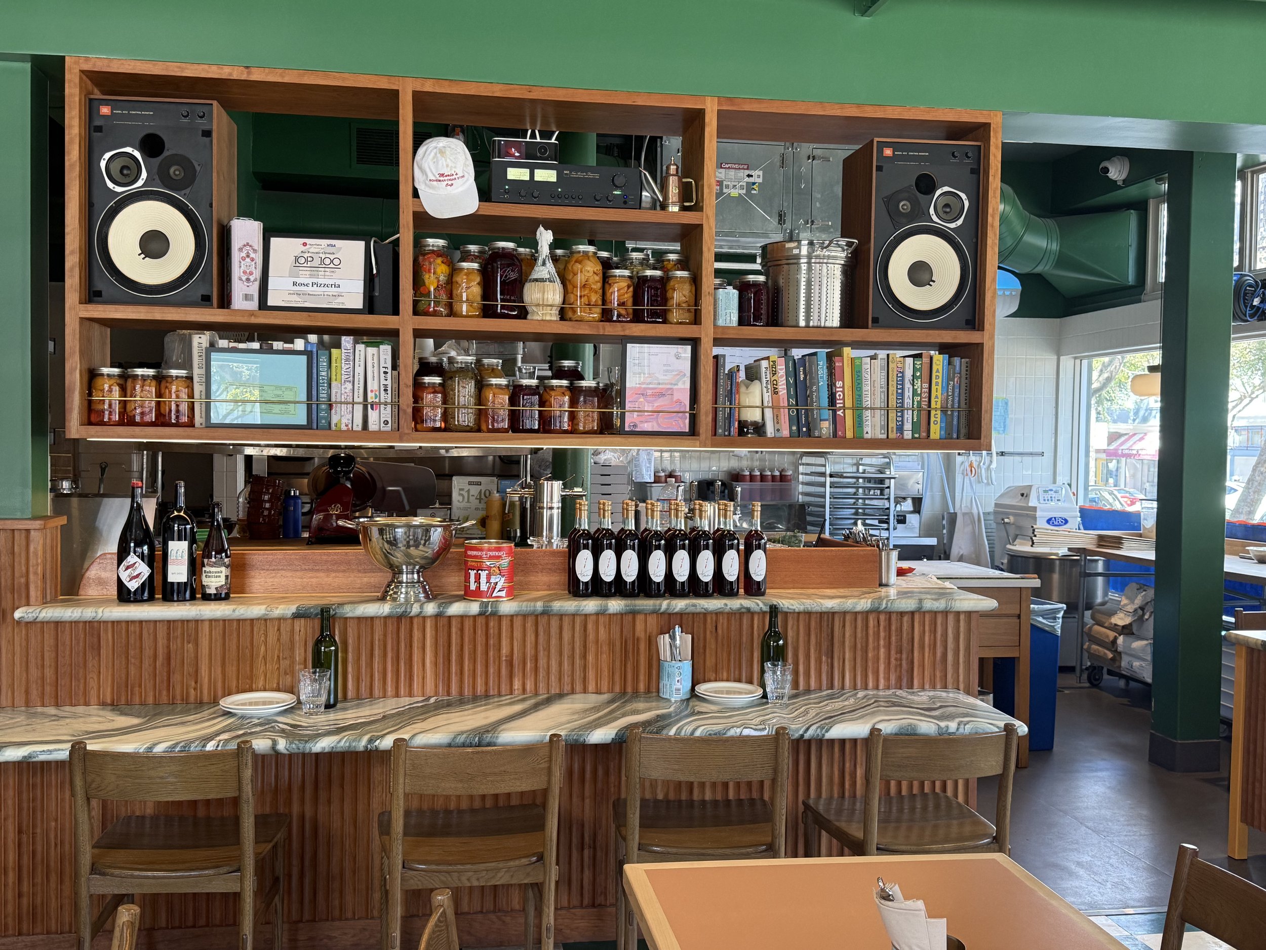 Interior of a restaurant or cafe with wooden shelves holding jars of preserved food, books, and audio equipment. There are bottles, glasses, and plates on the marble-topped counter in front. Green walls and windows provide natural light.