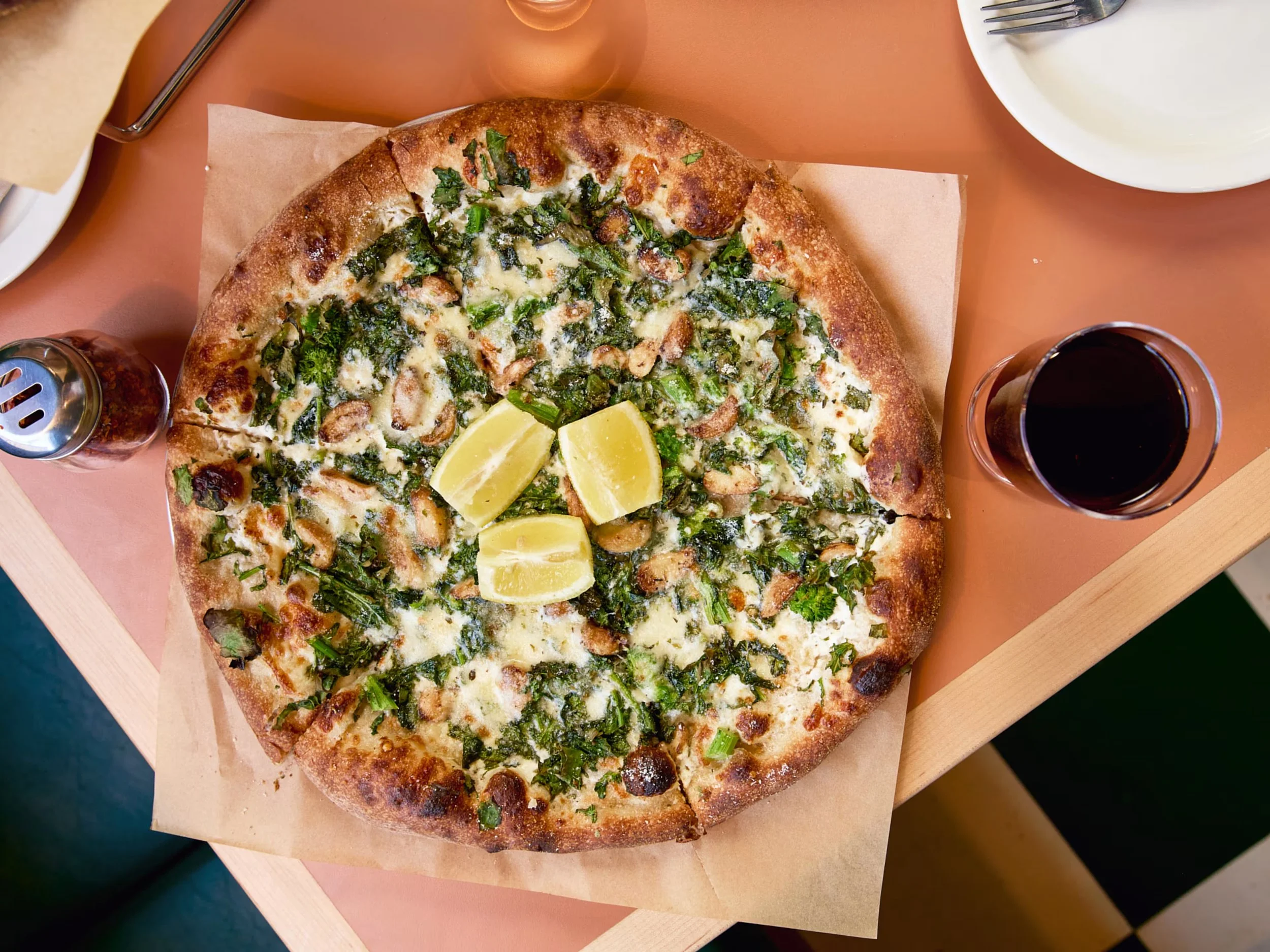 A pizza with green leafy vegetables, cheese, and lemon wedges on top, served on a wooden tray. There are plates and drinks nearby.