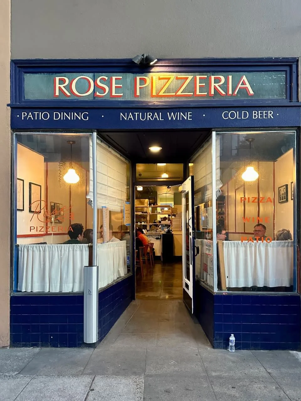 Exterior of Rose Pizzeria with blue sign and large glass windows showing inside seating, with customers dining, and a view into the kitchen area.