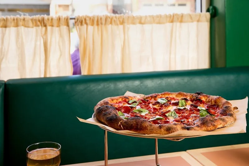 A pizza with basil leaves and pepperoni on a stand on a table in a restaurant, with a glass of drink nearby and cream-colored curtains in the background.