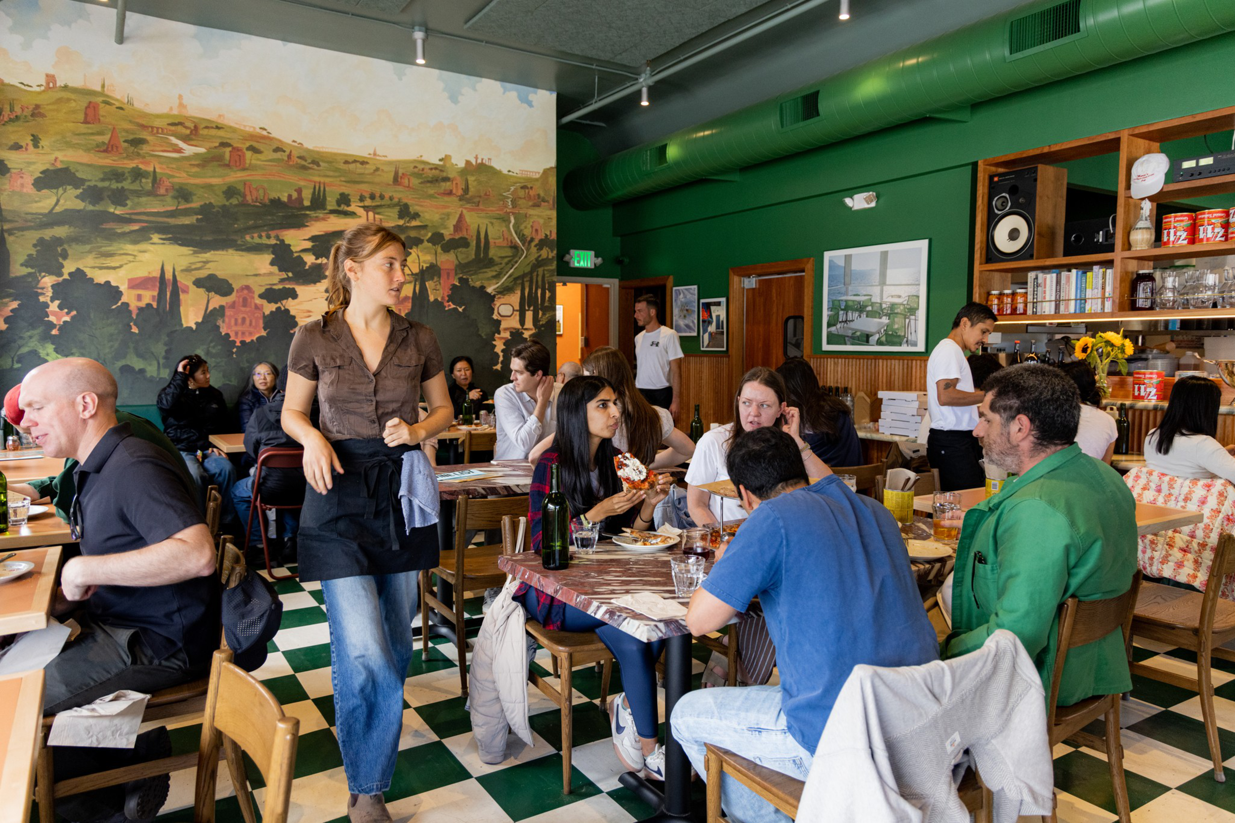 A busy restaurant with customers seated at tables and a waitress standing, holding a menu. The restaurant has a large wall mural depicting a scenic countryside landscape, and green walls with framed pictures. The atmosphere is lively with people dining and talking.
