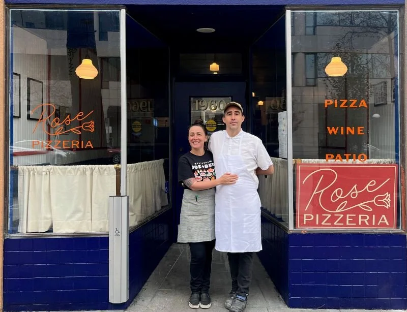 A woman and a man standing outside Rose Pizzeria, smiling. The woman is wearing a black T-shirt and a gray apron, and the man is in a white chef's coat and cap. The restaurant has signs indicating it serves pizza, wine, and has a patio. 
