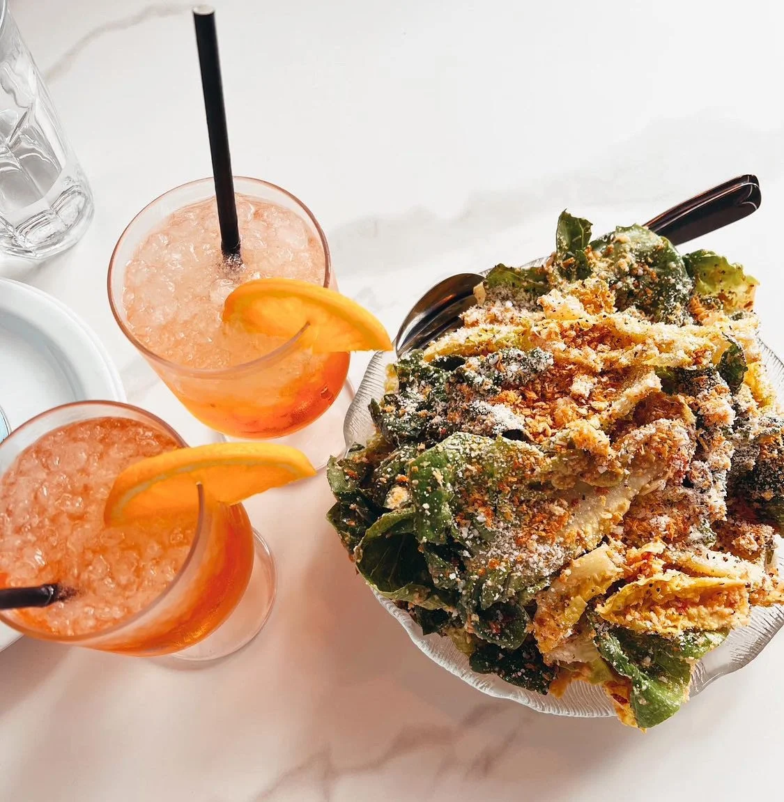 Two pink drinks with black straws and orange slices, and a large salad topped with cheese and croutons on a white table.