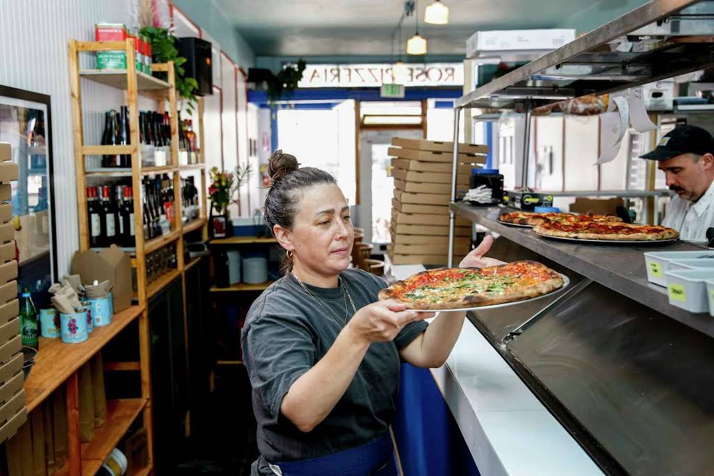 A woman in black shirt holding a pizza in a pizzeria kitchen, with a man behind a counter preparing pizza, shelves with wine and supplies, and pizza boxes nearby.