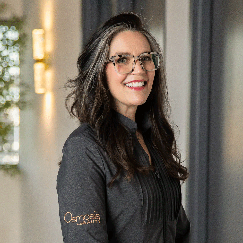 Smiling woman with long dark hair, glasses with animal print frames, wearing a dark gray zip-up jacket with 'Osmosis Beauty' logo, standing indoors with blurred background.