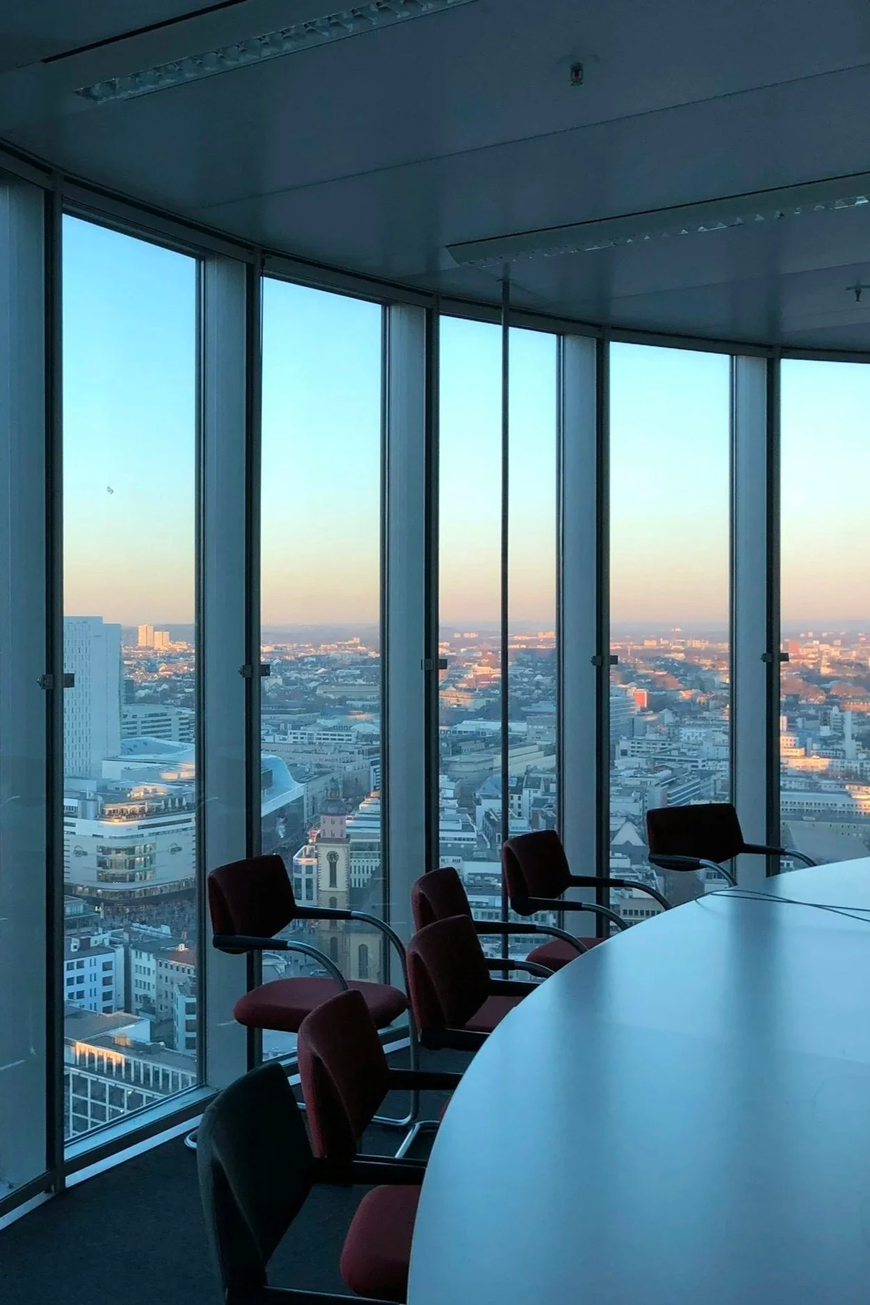 A conference room with a large, round table and red office chairs, glass walls displaying a city skyline during sunrise or sunset, and ceiling lights.