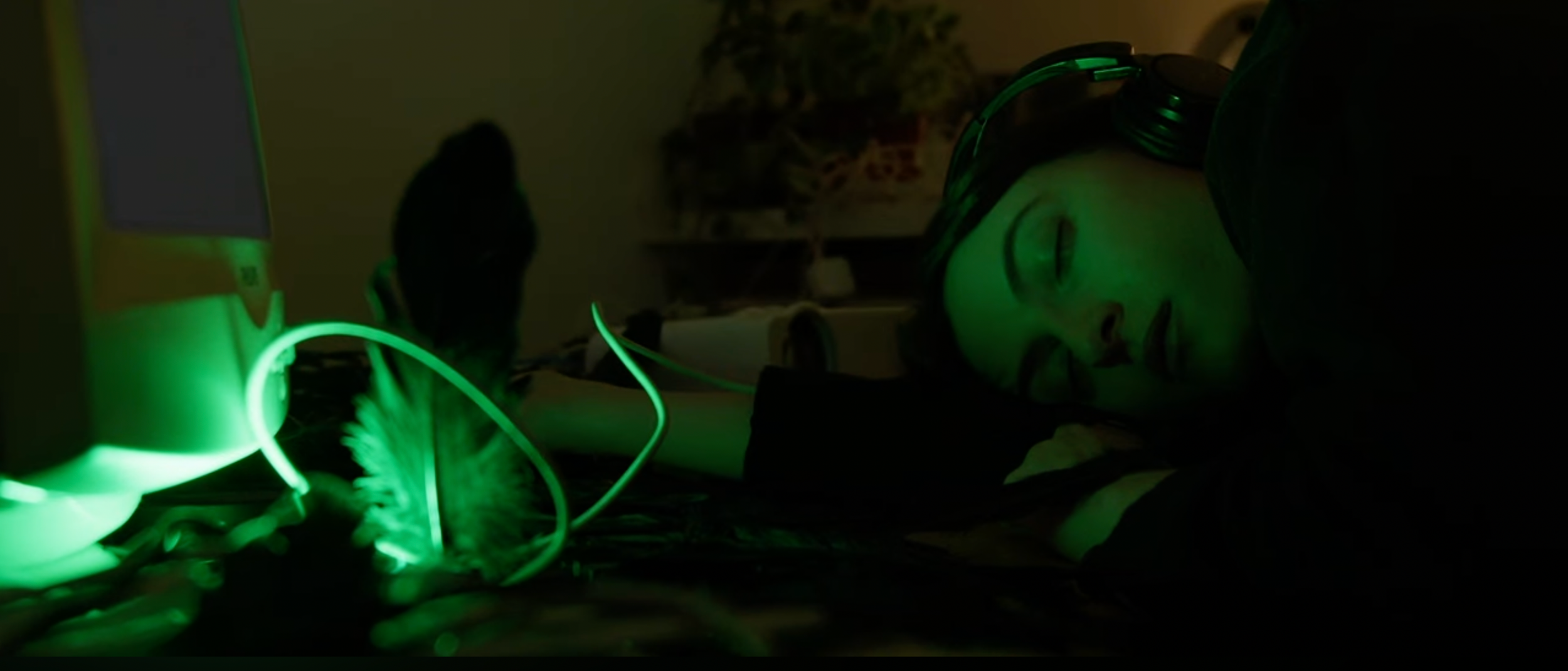 Person sleeping on a desk with headphones, illuminated by green glow from electronic devices.