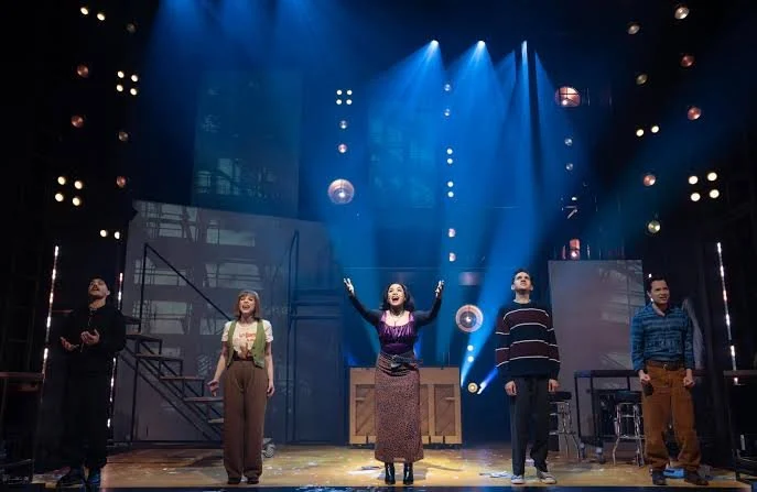 Five actors on stage performing in a theater with bright stage lighting and decorative hanging lights.