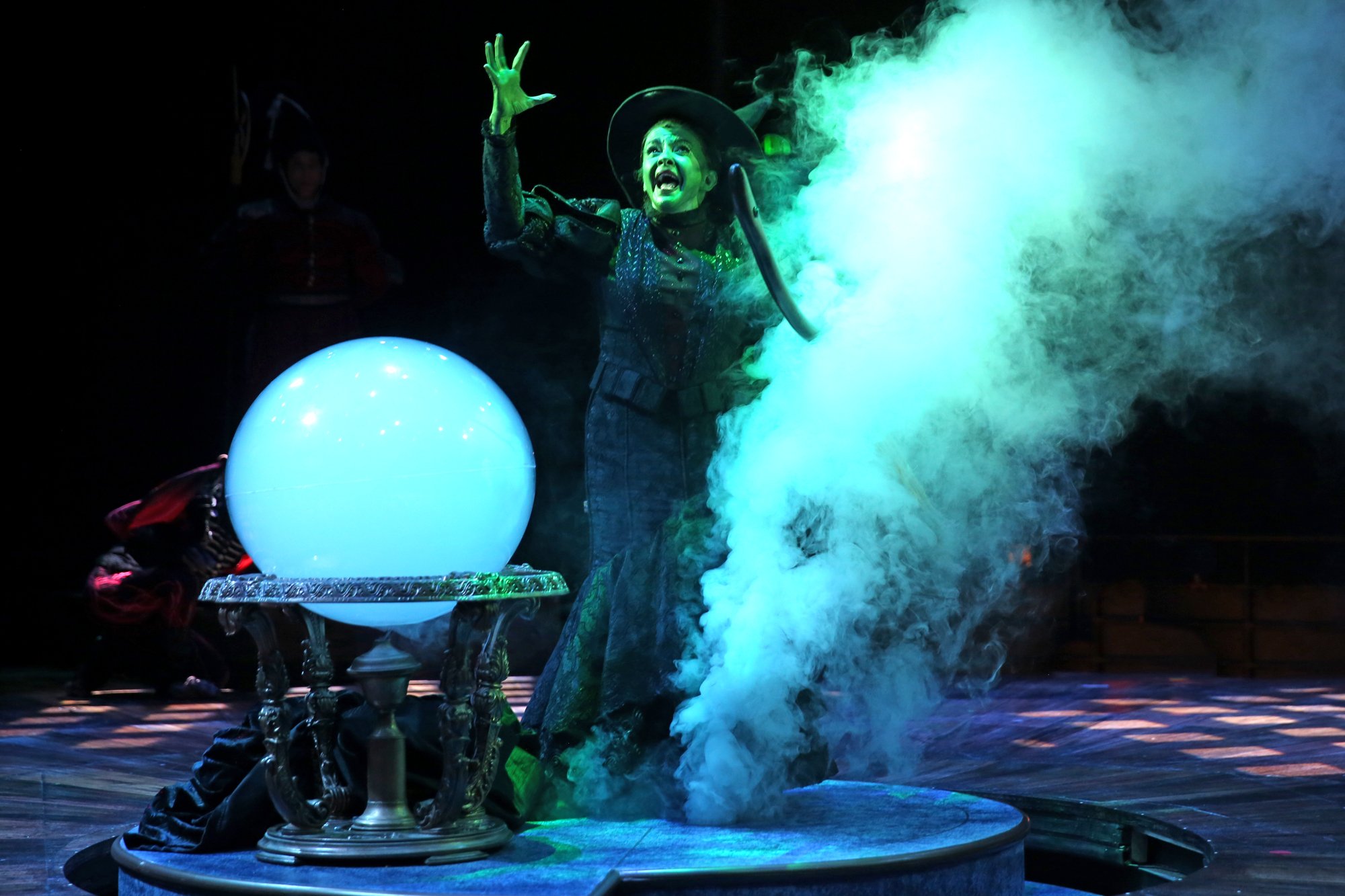 A woman dressed as a witch performing on stage with smoke and a glowing blue orb on a table in front of her.