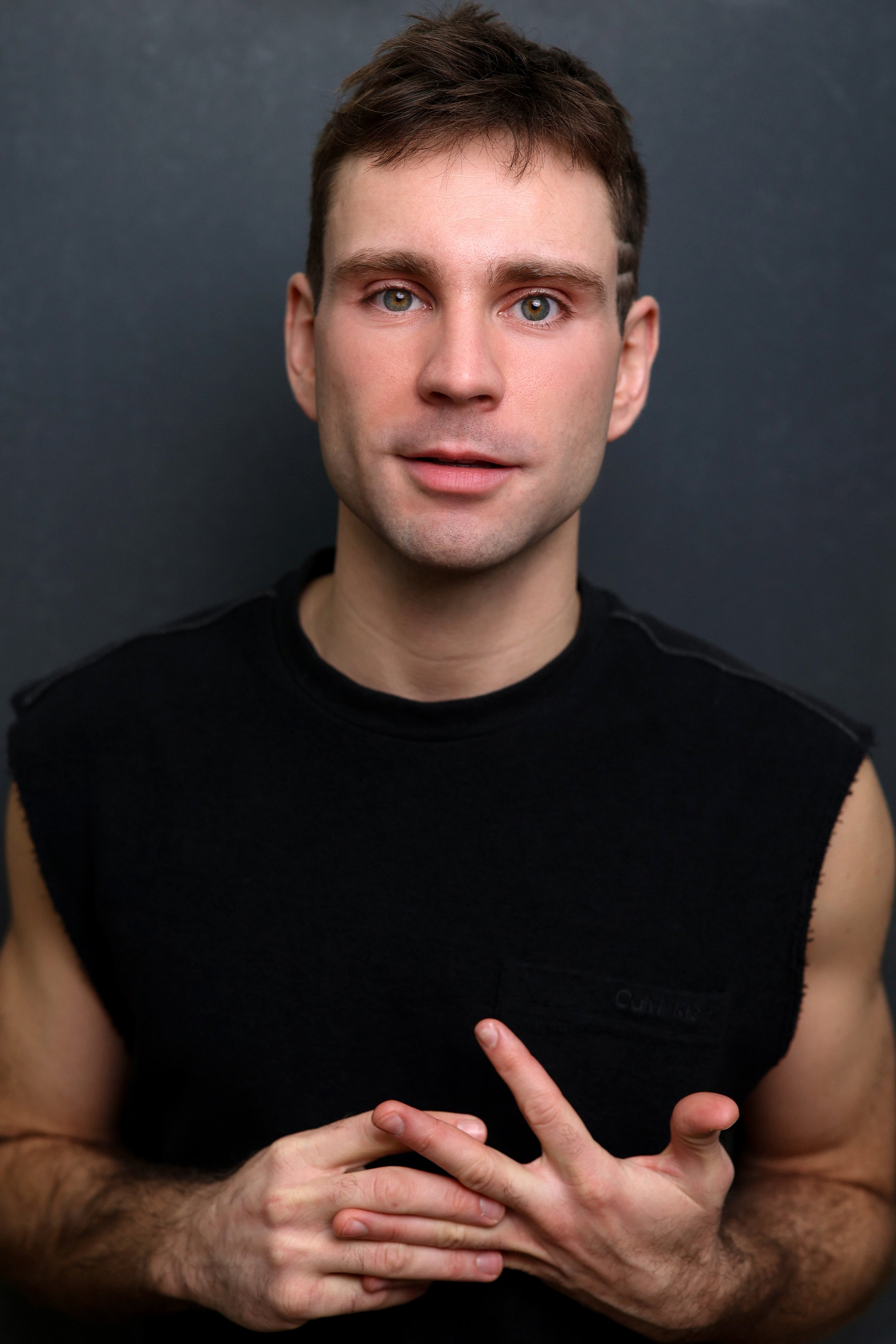 Close-up of a man pointing to his chest with his right hand against a dark background. He has short brown hair, light green eyes, and is wearing a sleeveless black shirt.